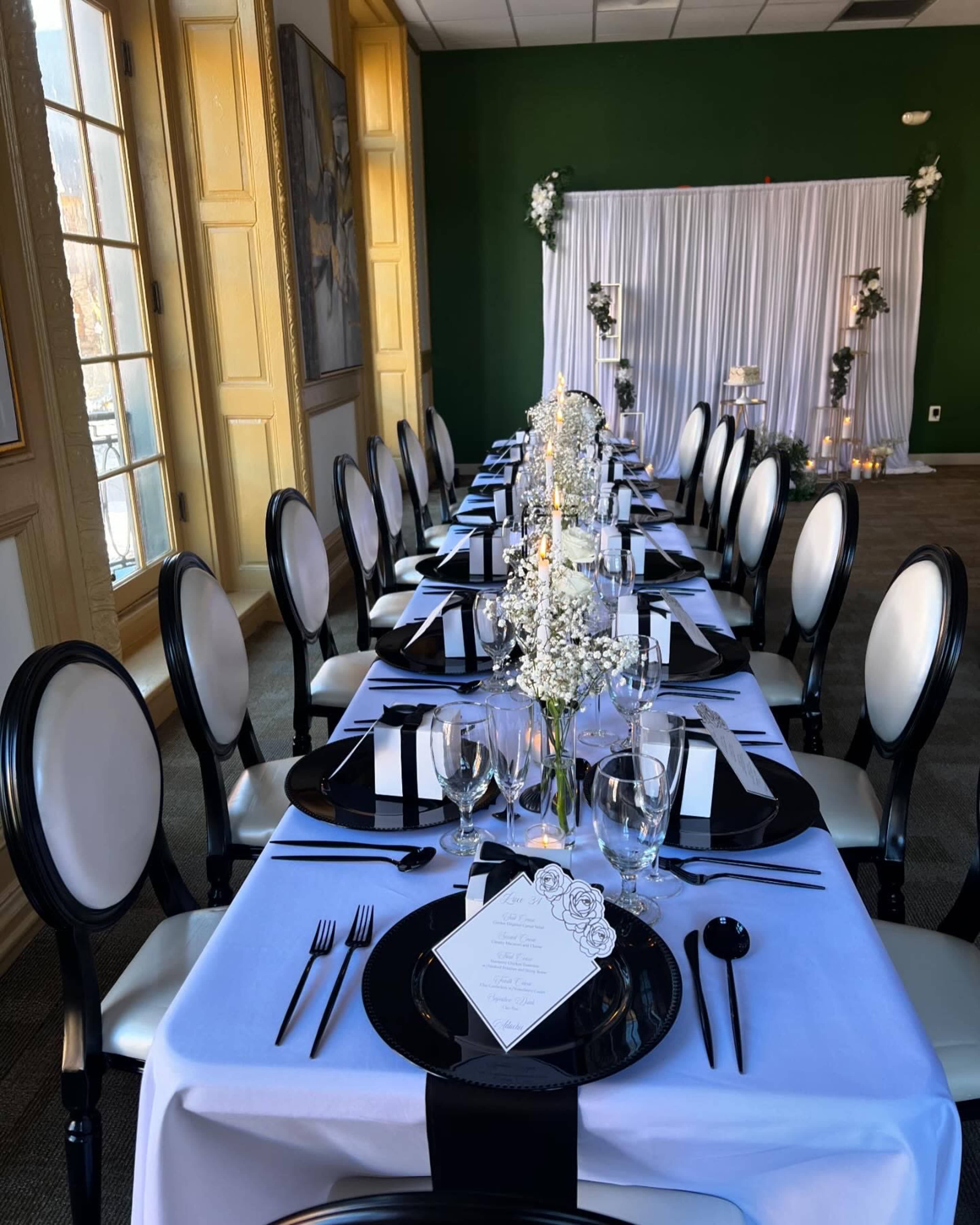 A long dining table is set with black and white tableware, adorned with flowers and candles, in a room with green walls and large windows.
