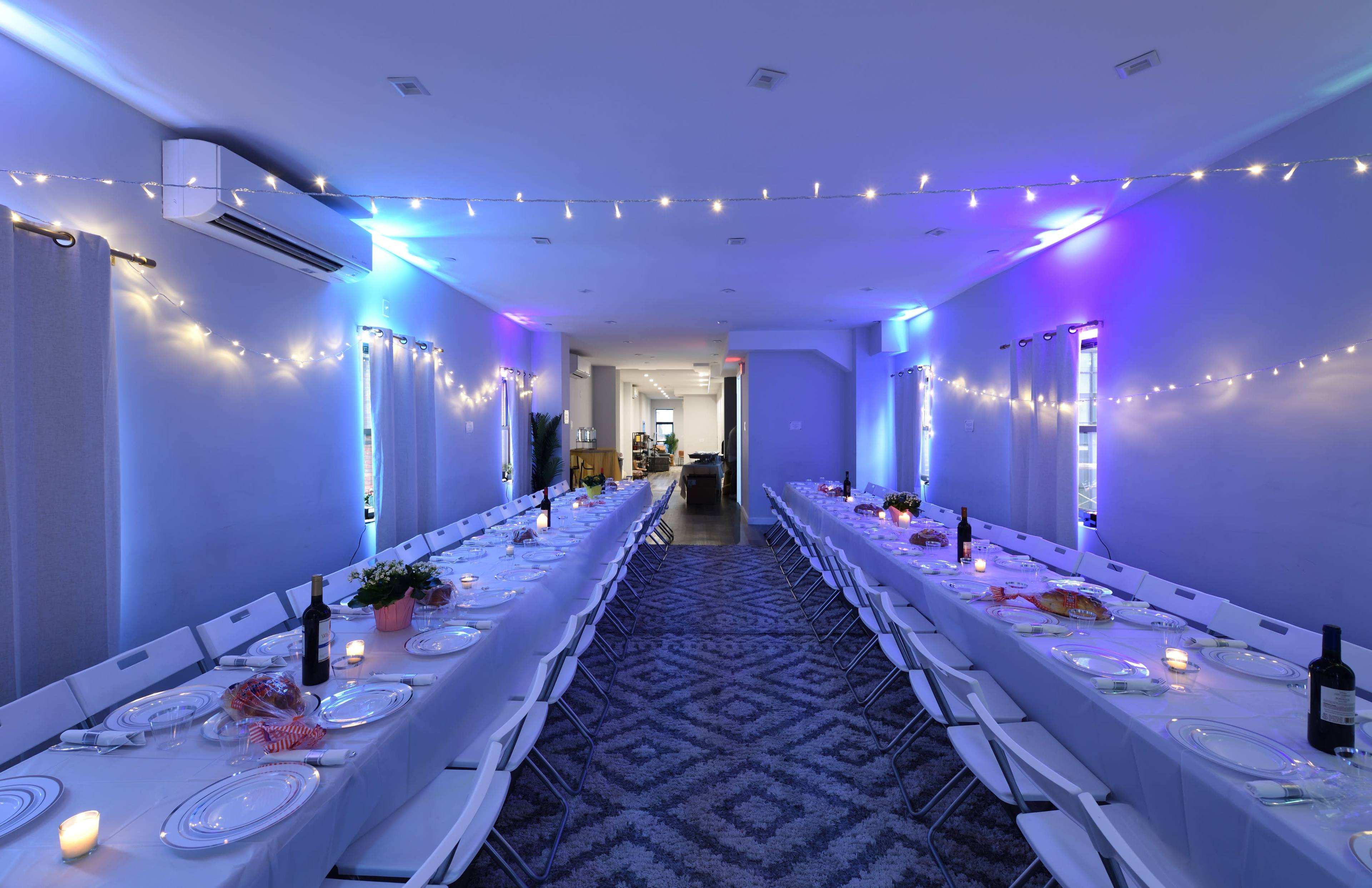 A long, rectangular dining table set for a meal is illuminated by colorful lights and adorned with candles in a spacious, modern room.