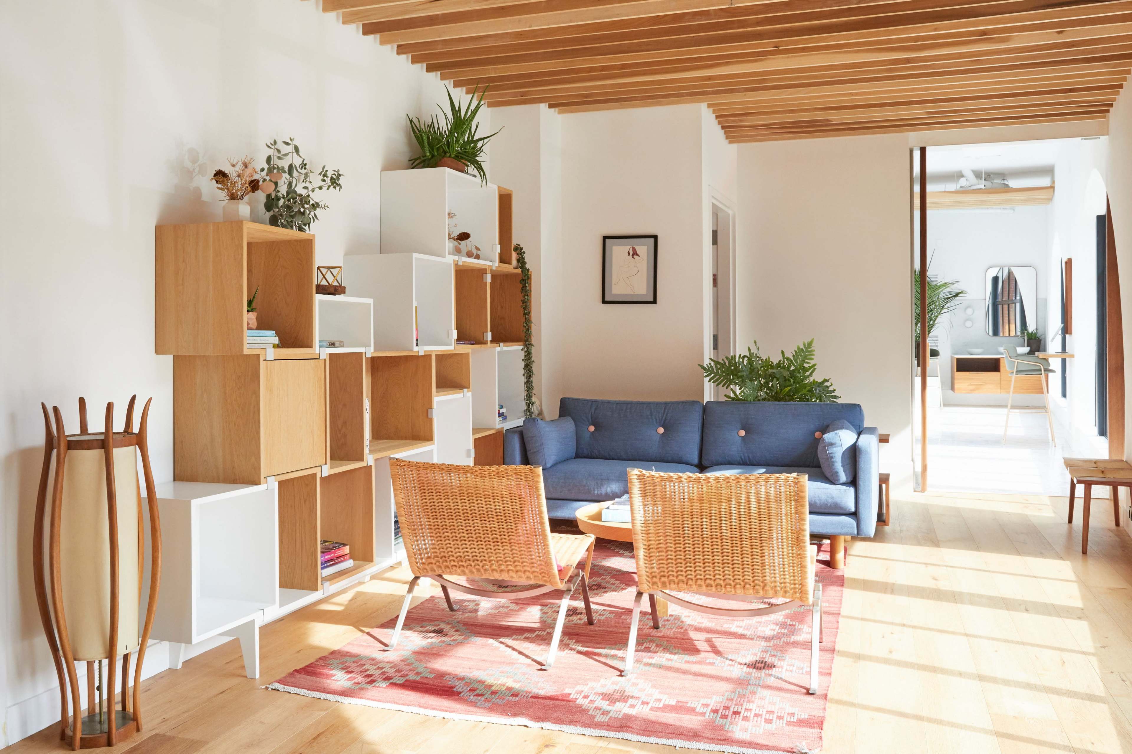 A bright, modern living room with a blue sofa, two rattan chairs, a coffee table, and wooden shelving against a white wall.