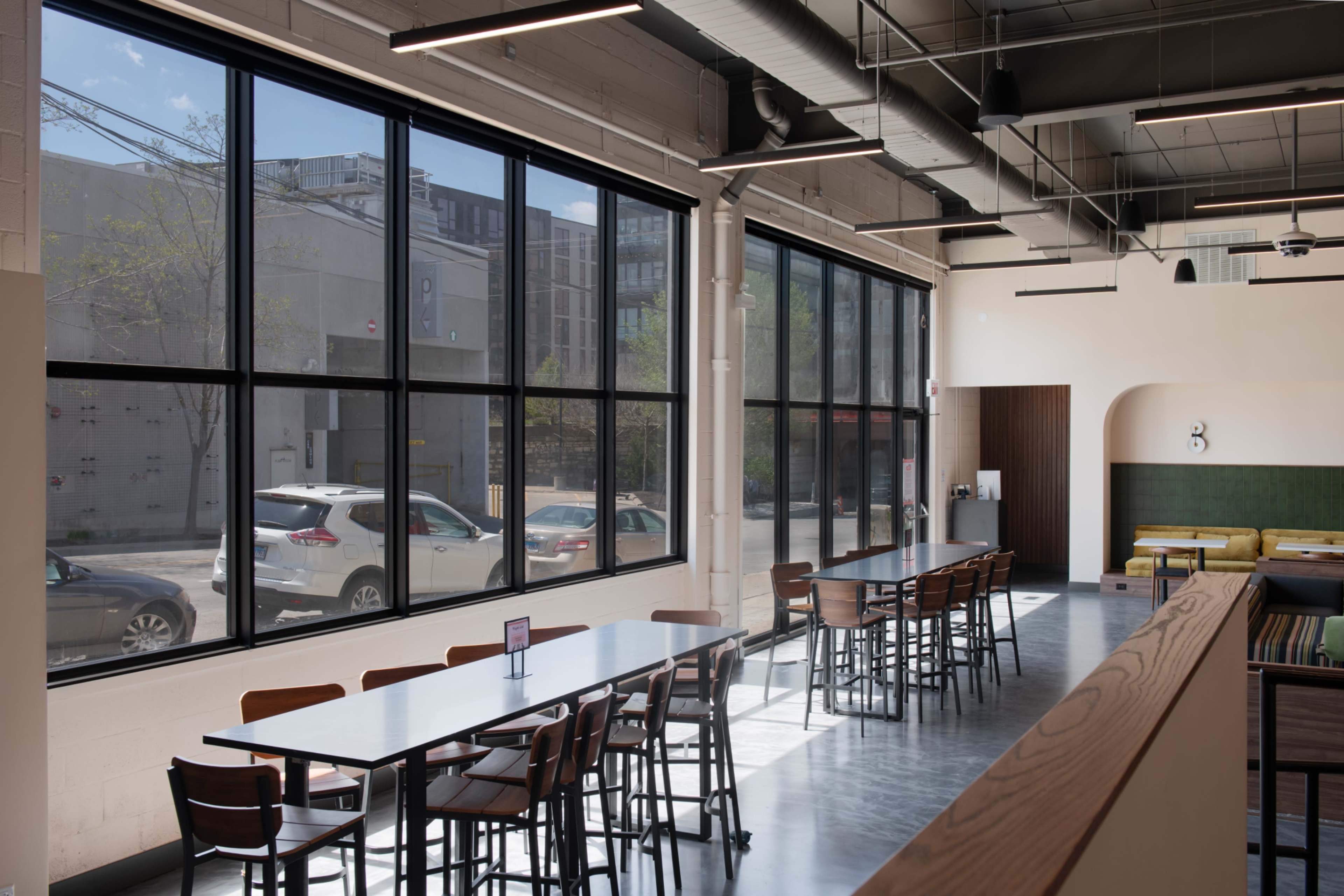 The image shows a spacious, modern café interior with large windows allowing natural light to fill the room, featuring rows of tables and chairs along one side.