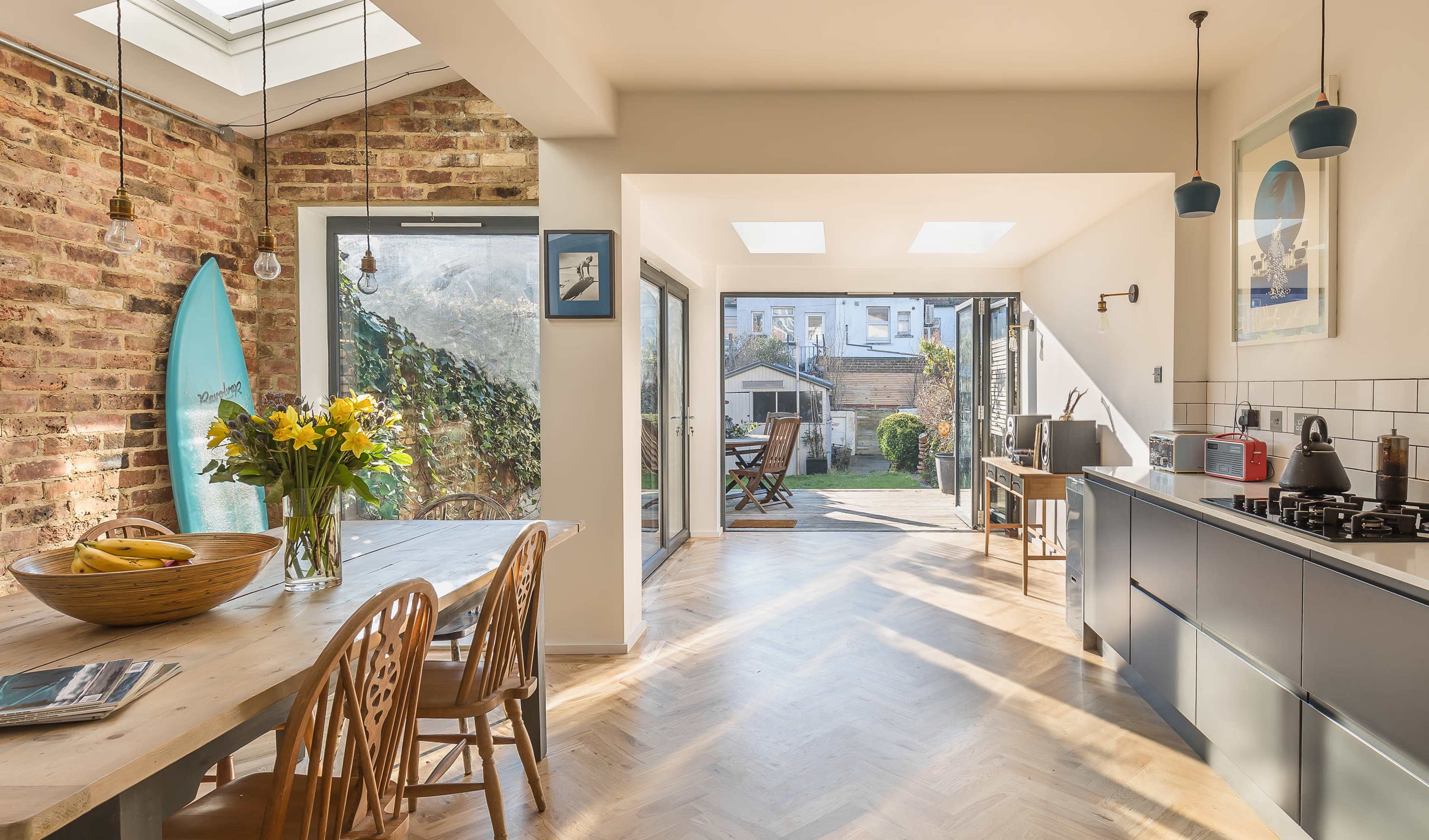 A modern kitchen and dining area features a wooden table with chairs, a bouquet of flowers, and large glass doors opening to a sunny garden with a surfboard leaning against the wall.