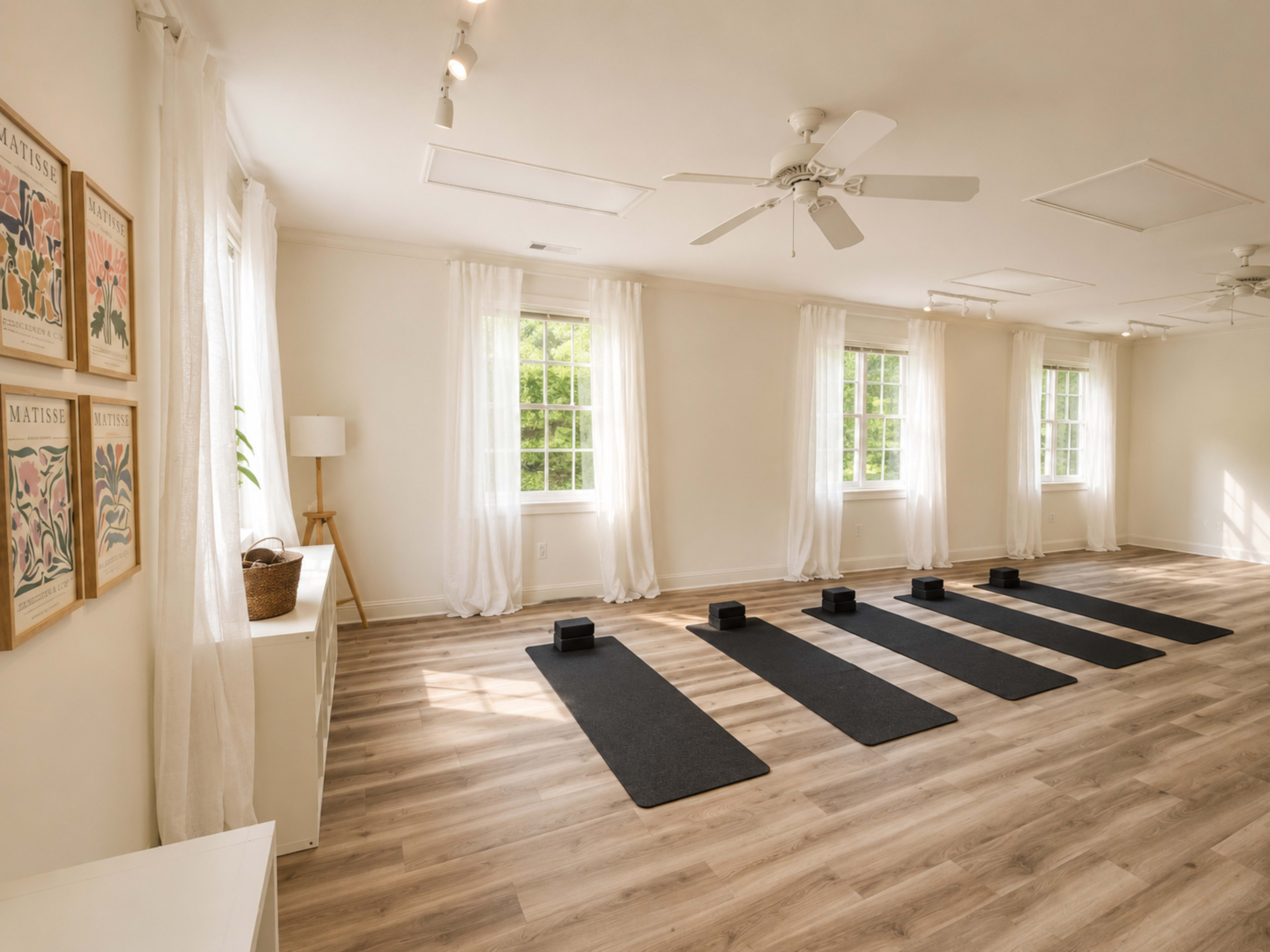 The image shows a bright yoga studio with four black mats arranged on a wooden floor, surrounded by large windows with sheer white curtains.