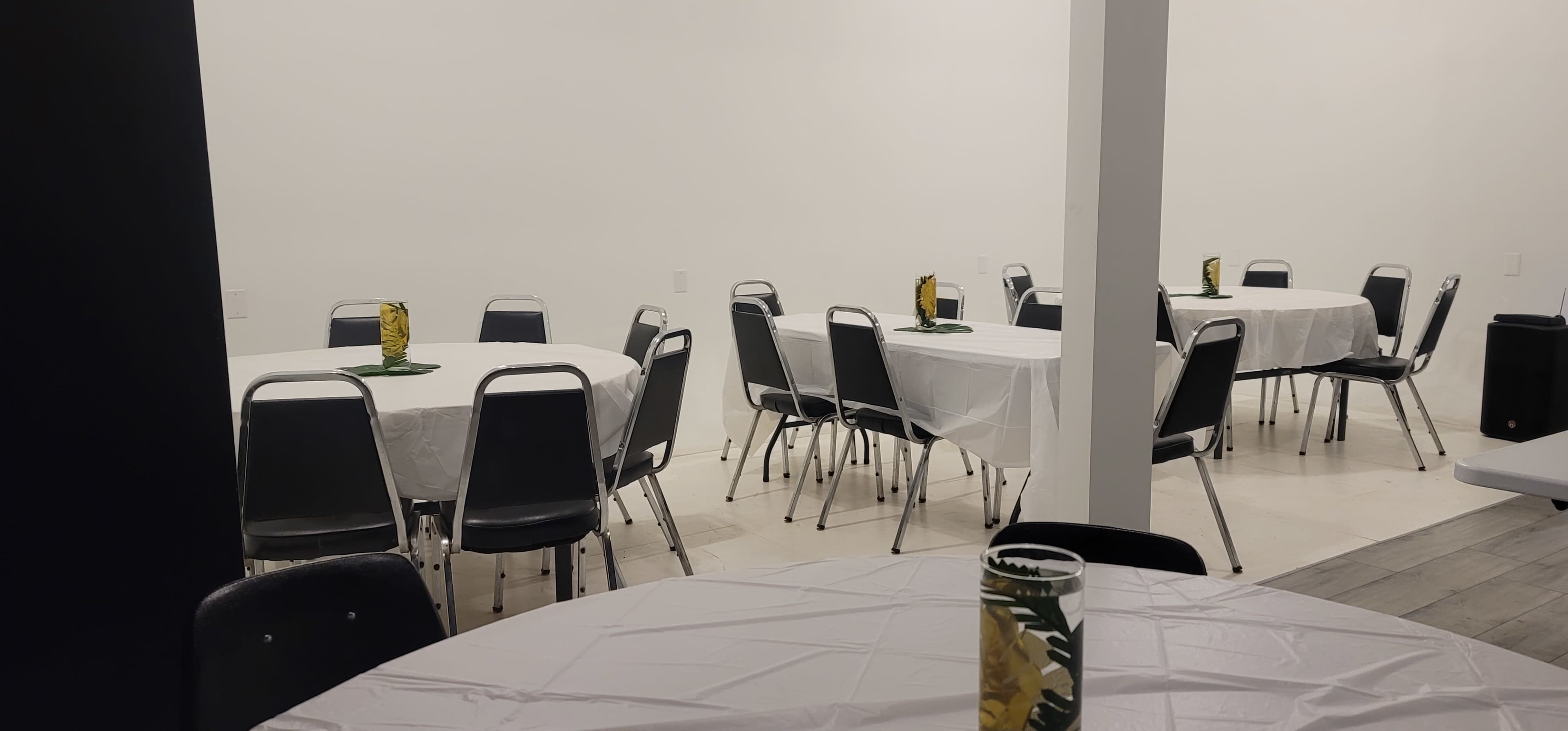 A spacious room arranged with several tables covered in white tablecloths and black chairs, each decorated with green leaves and glassware.