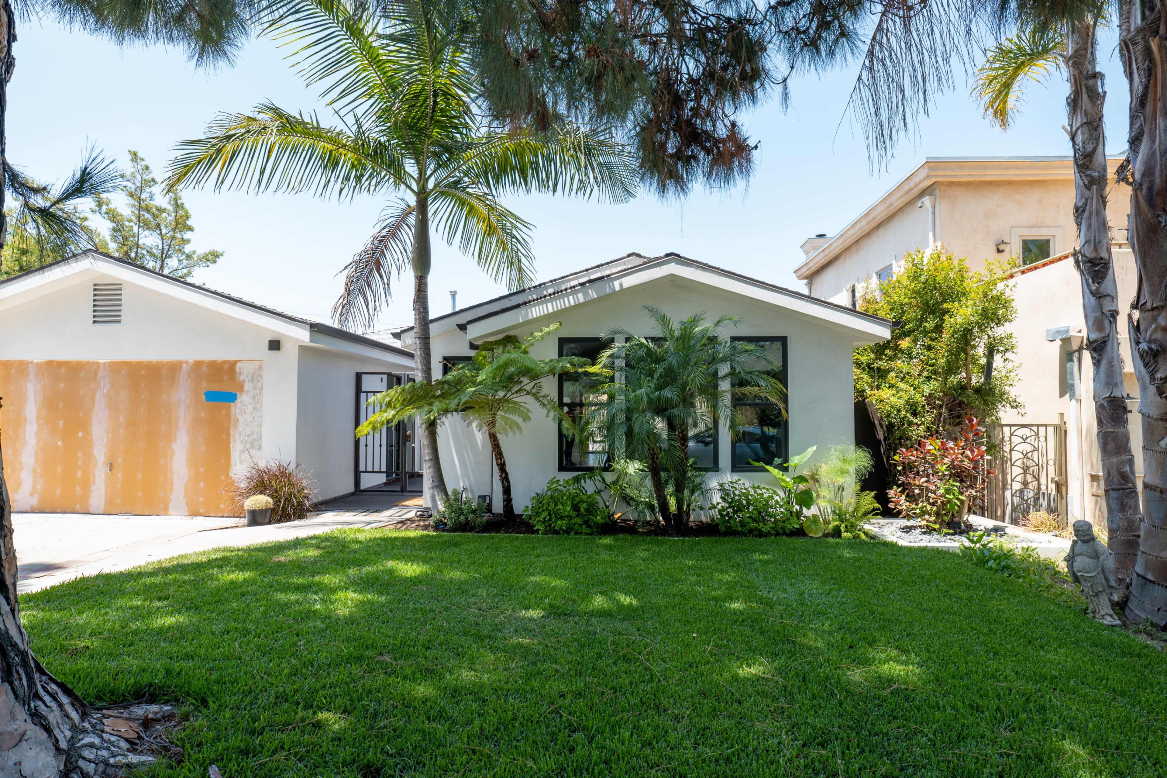 A single-story house with a manicured lawn, palm trees, and tropical plants is shown beside a garage with a yellow exterior.