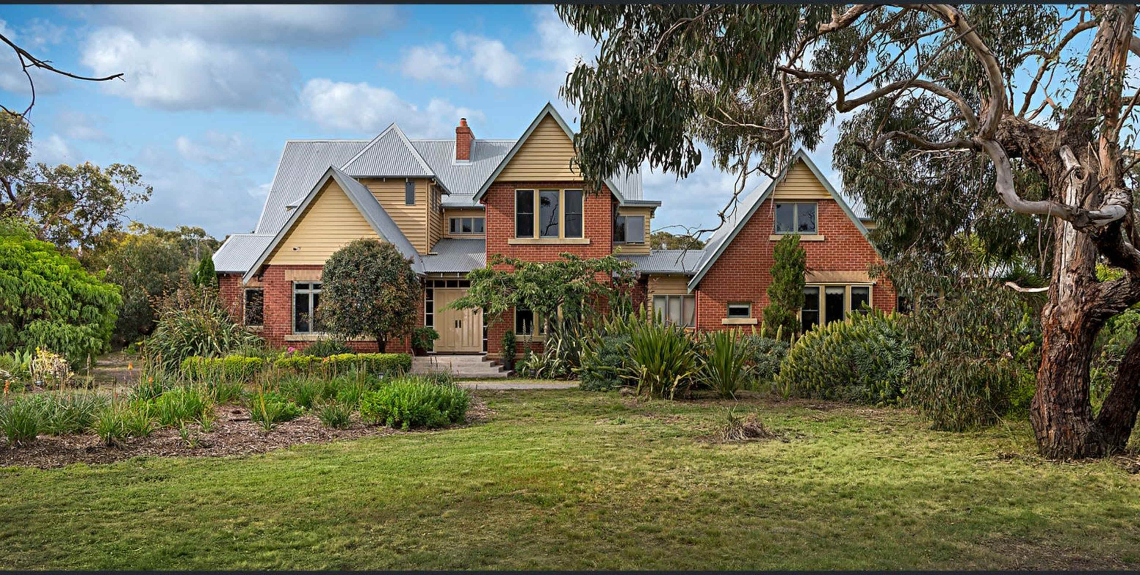 A large two-story house with a combination of brick and wooden siding is surrounded by a well-maintained garden and trees.