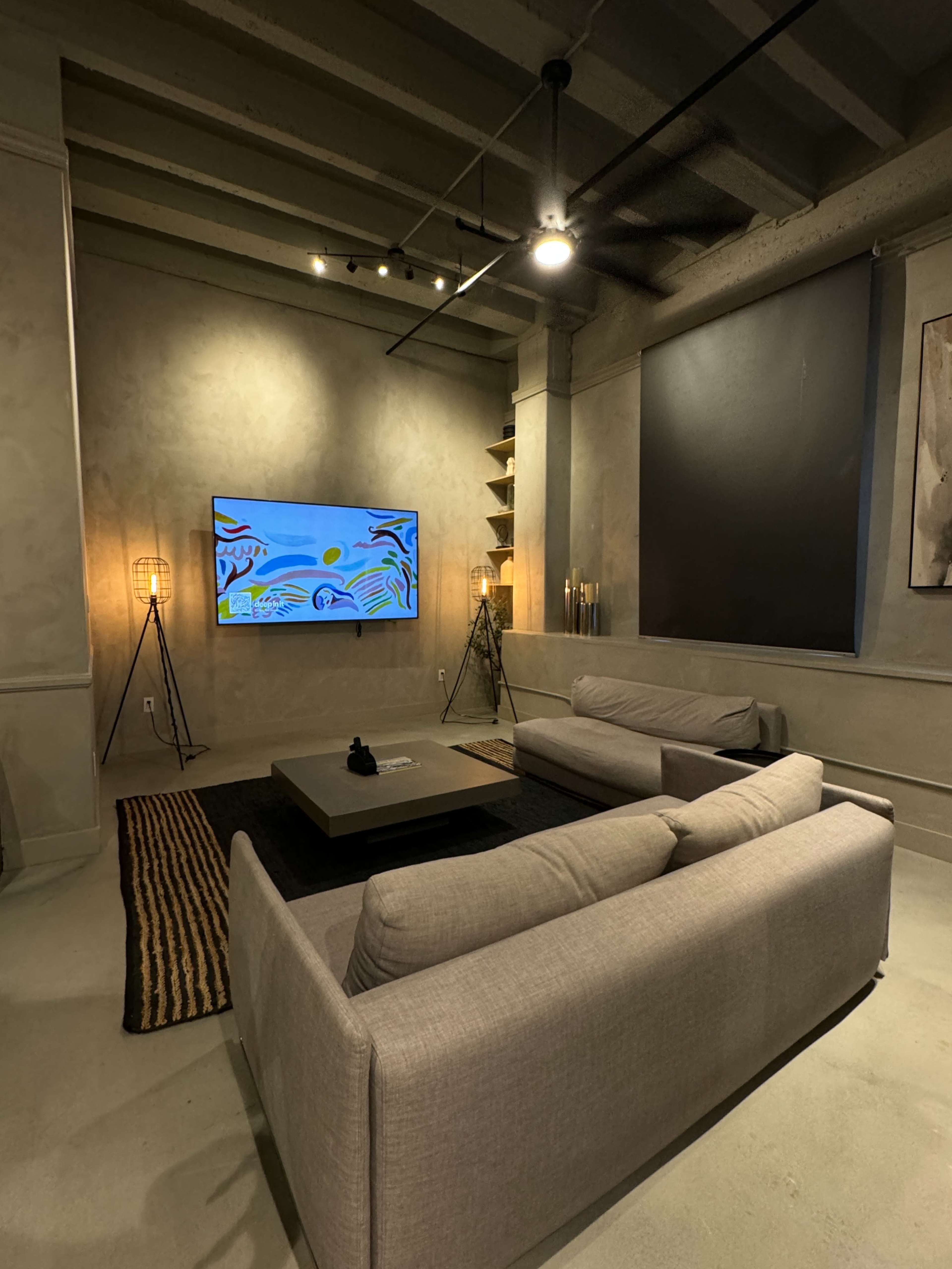 The image shows a modern living room with two gray sofas facing a large wall-mounted television, accented by industrial lighting and shelves.
