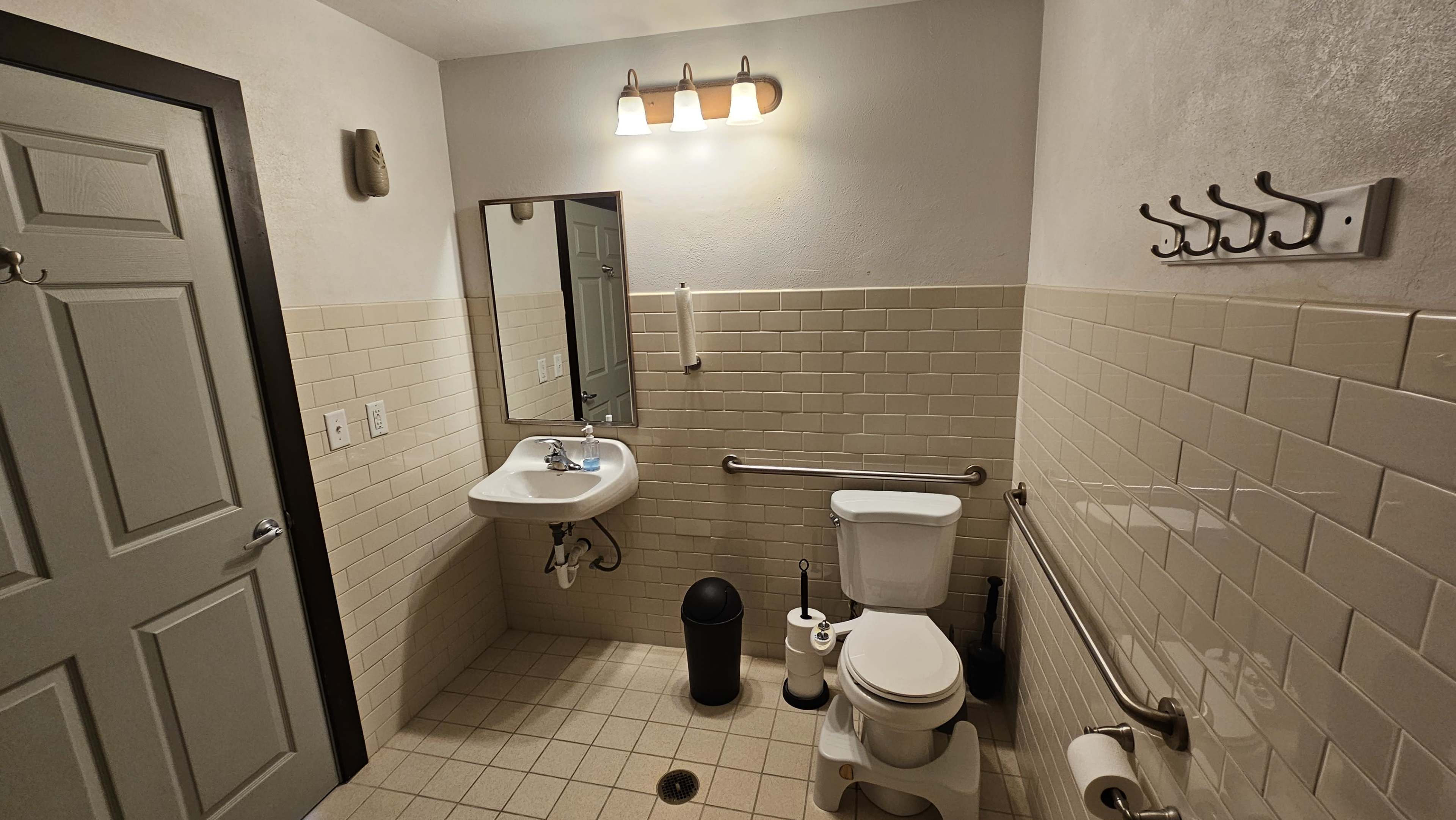 The image shows a small bathroom featuring a toilet, a wall-mounted sink with a mirror, and grab bars along the wall.