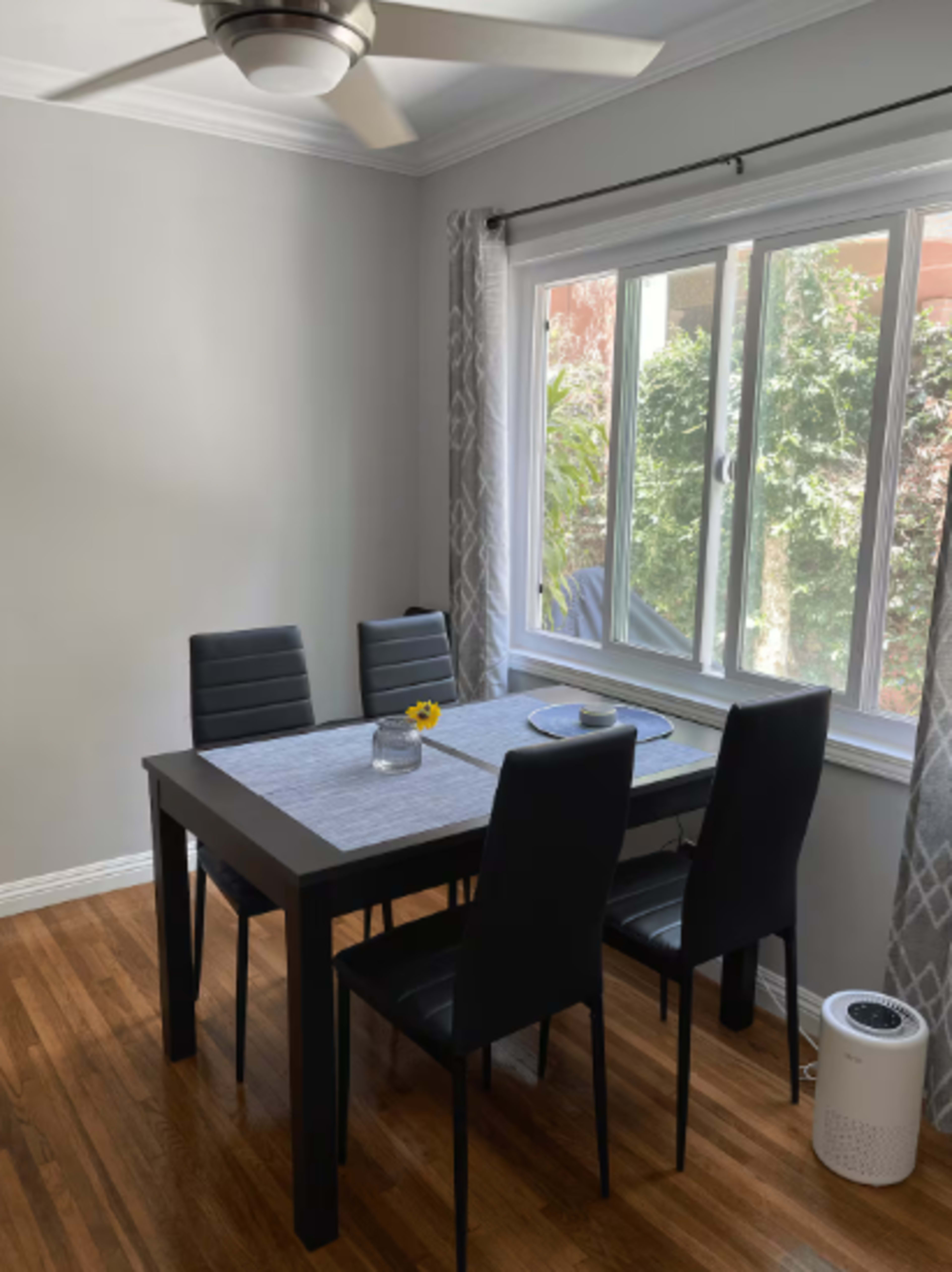 A black dining table with four chairs is placed in a well-lit room beside a large window.