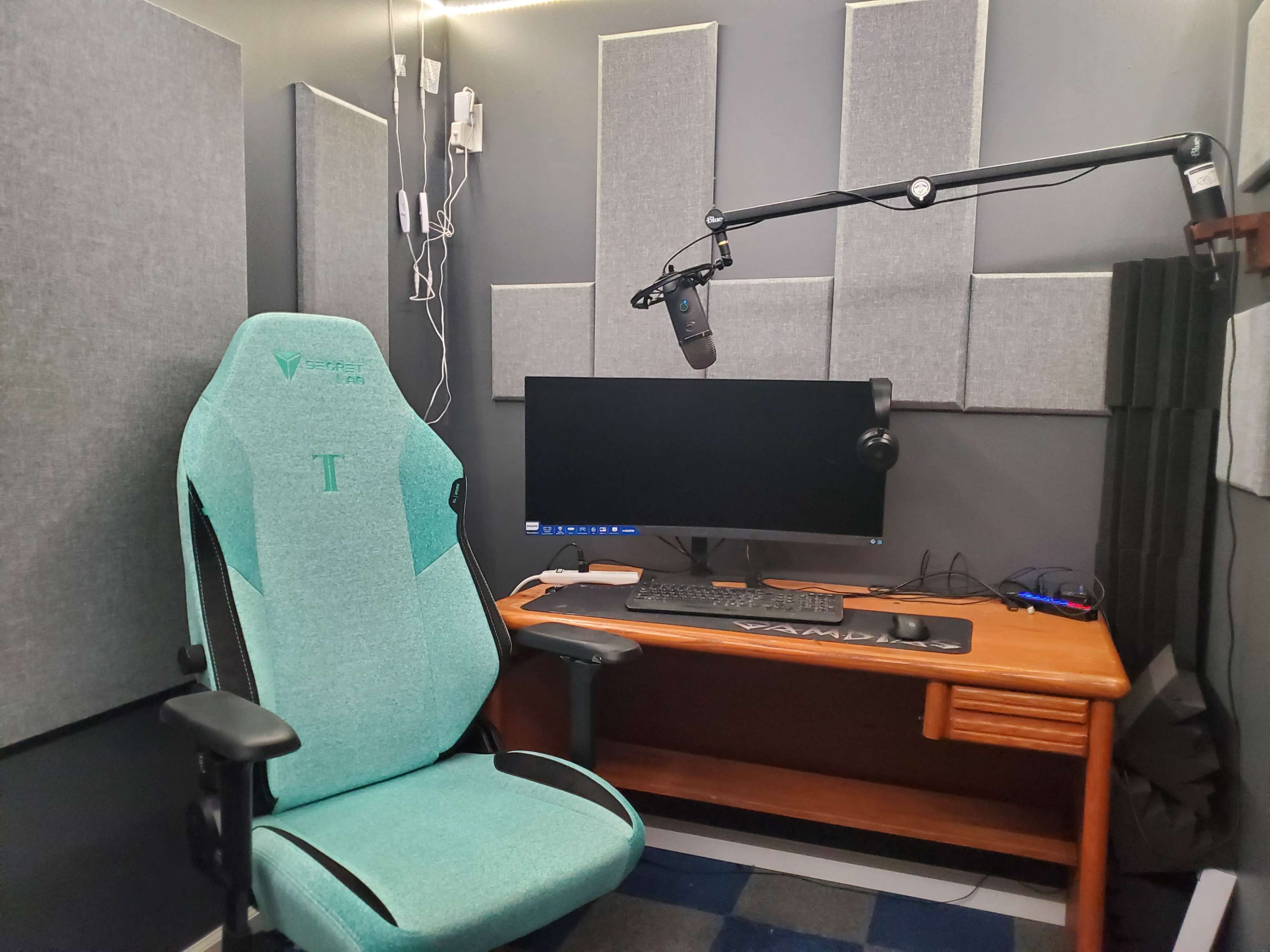 A recording setup featuring a teal office chair, a wooden desk with a computer monitor, a microphone on an adjustable arm, and soundproofing panels on the walls.