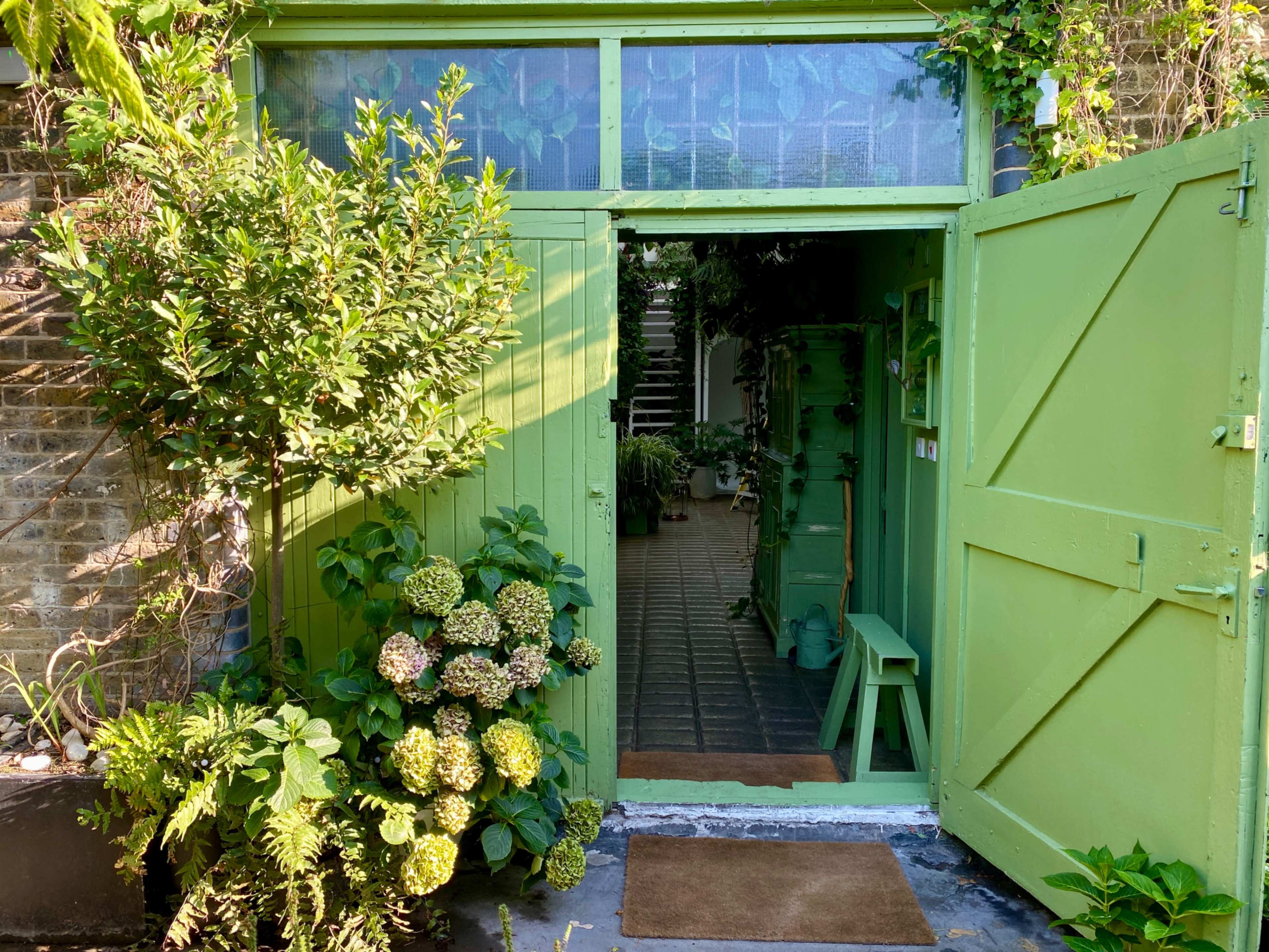 A bright green double door opens into an indoor space surrounded by various plants and greenery.