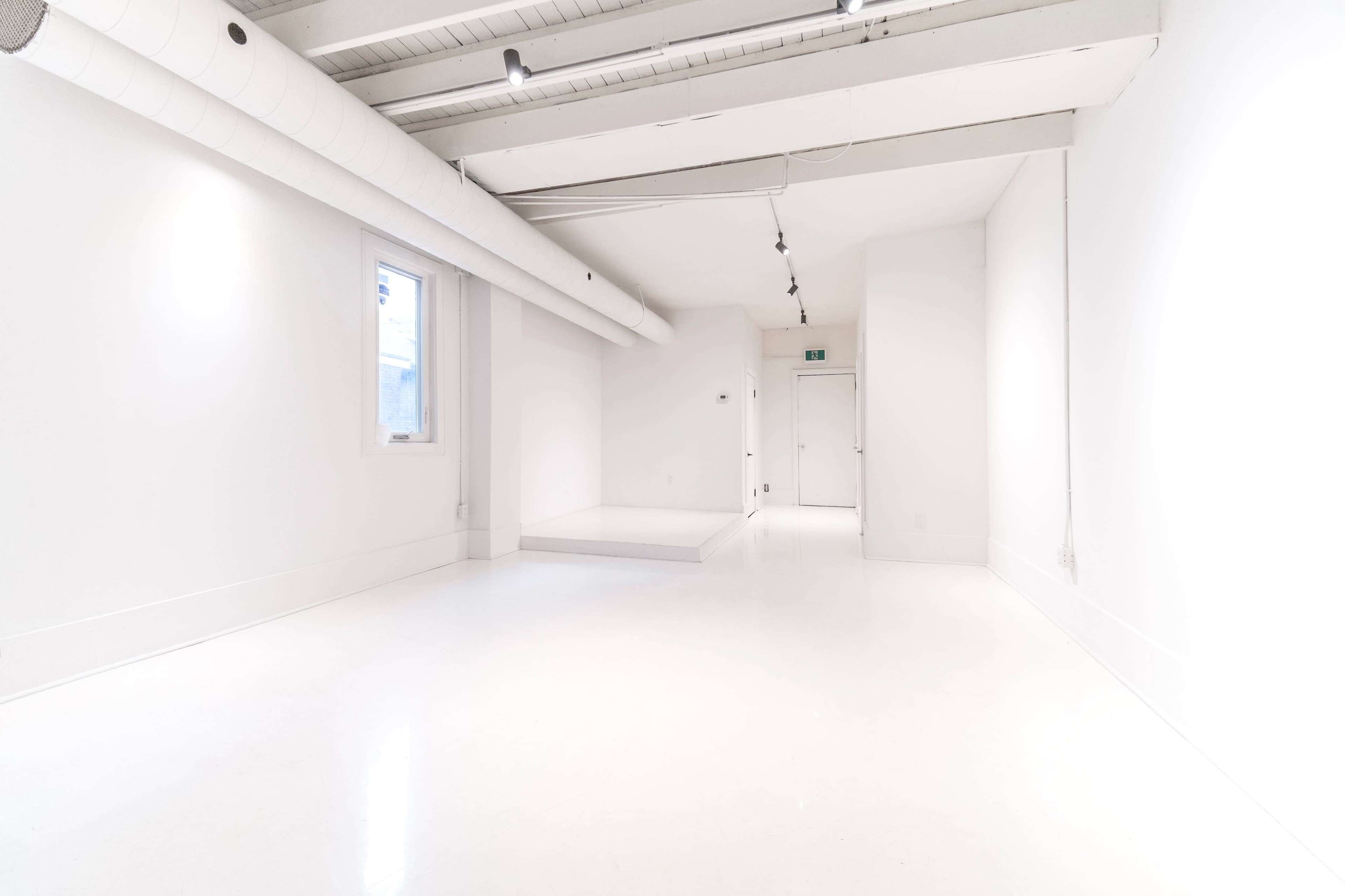 A spacious, well-lit white room with a smooth floor, a window on the left, and a door at the far end.