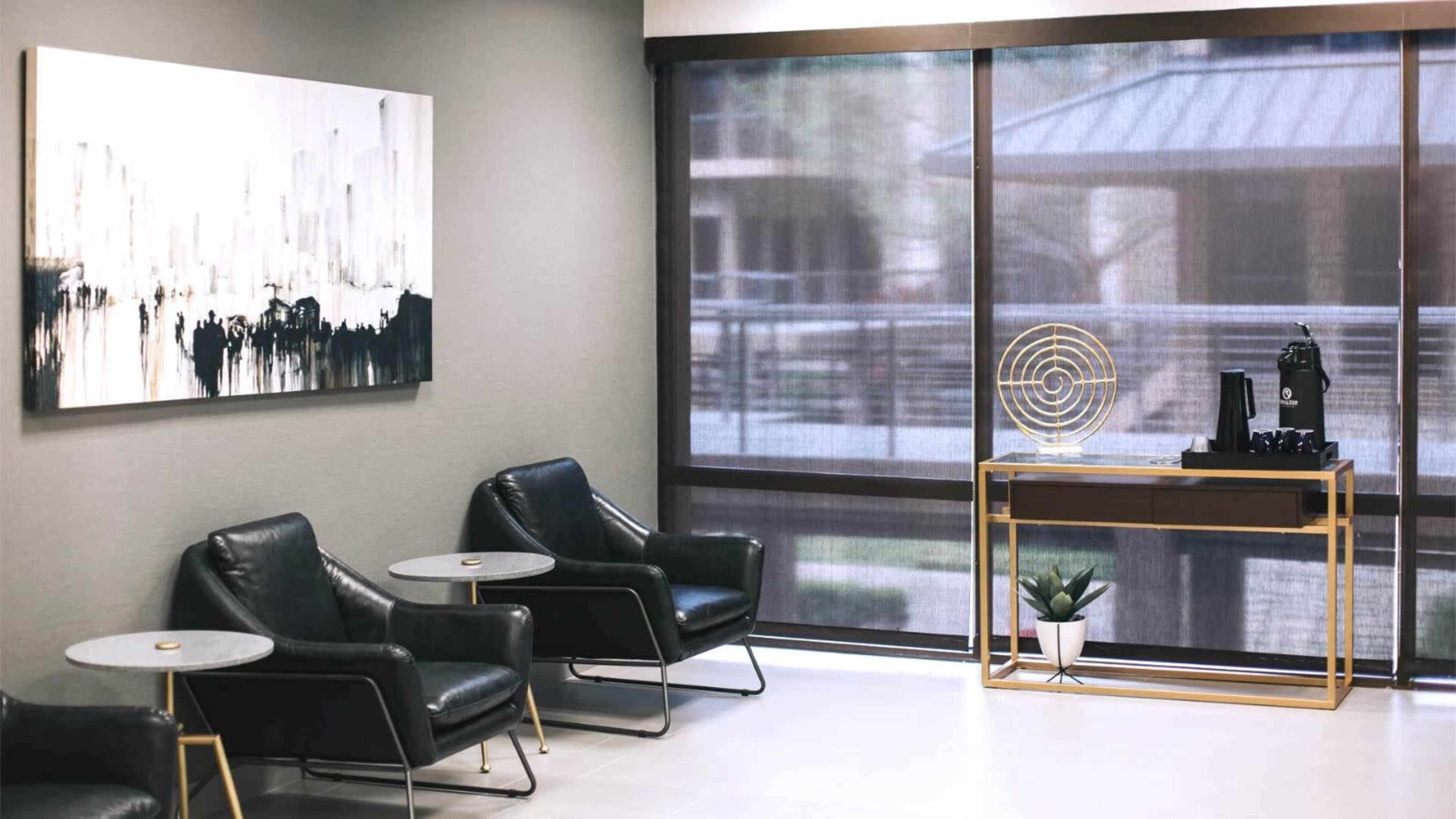 The image shows a modern waiting area with black leather chairs, small round tables, and a console table displaying a coffee machine and decor, against a backdrop of large windows.