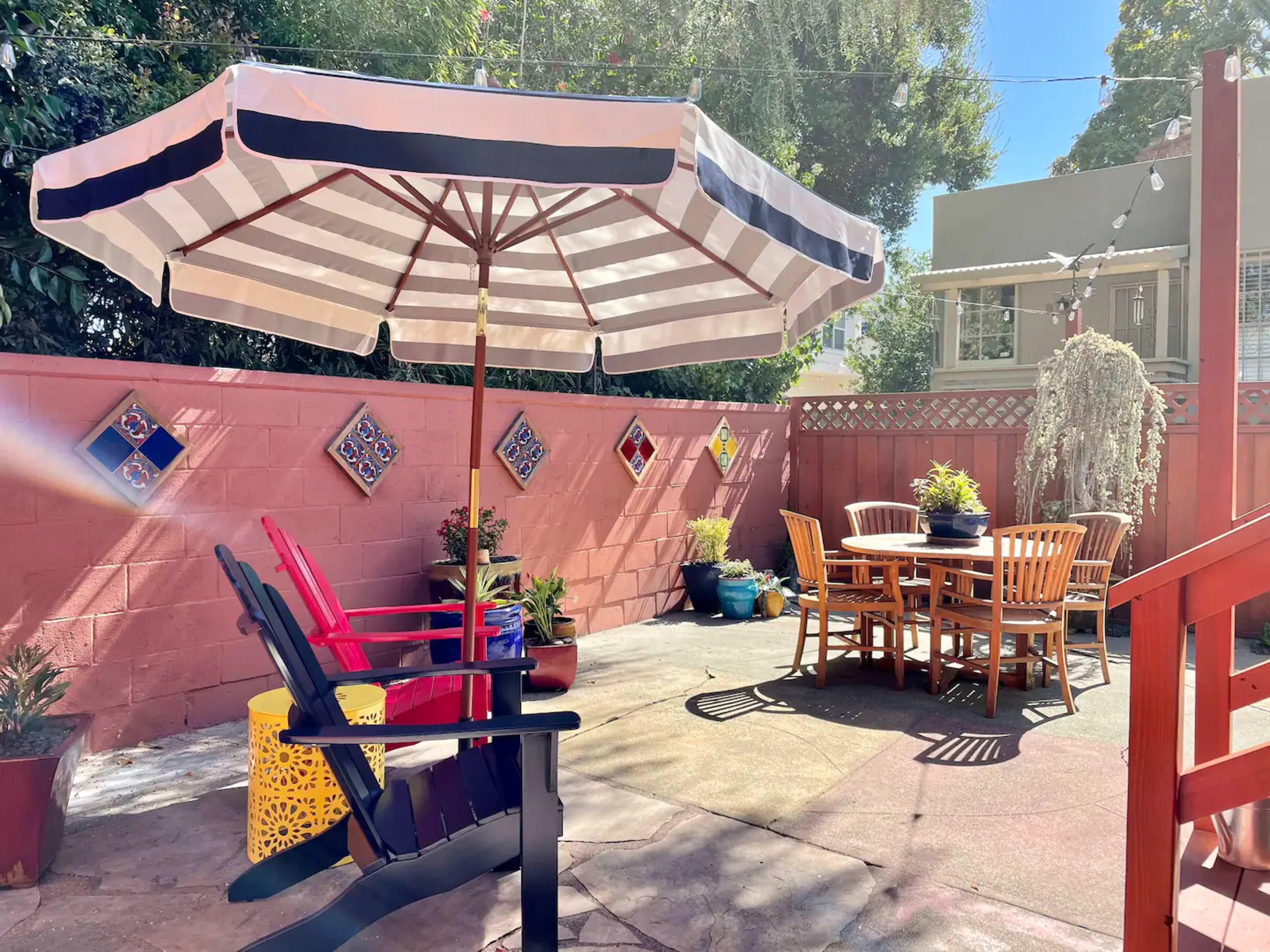 The scene features a colorful outdoor patio with a large striped umbrella, wooden dining table, and two adirondack chairs surrounded by potted plants and a decorative wall.