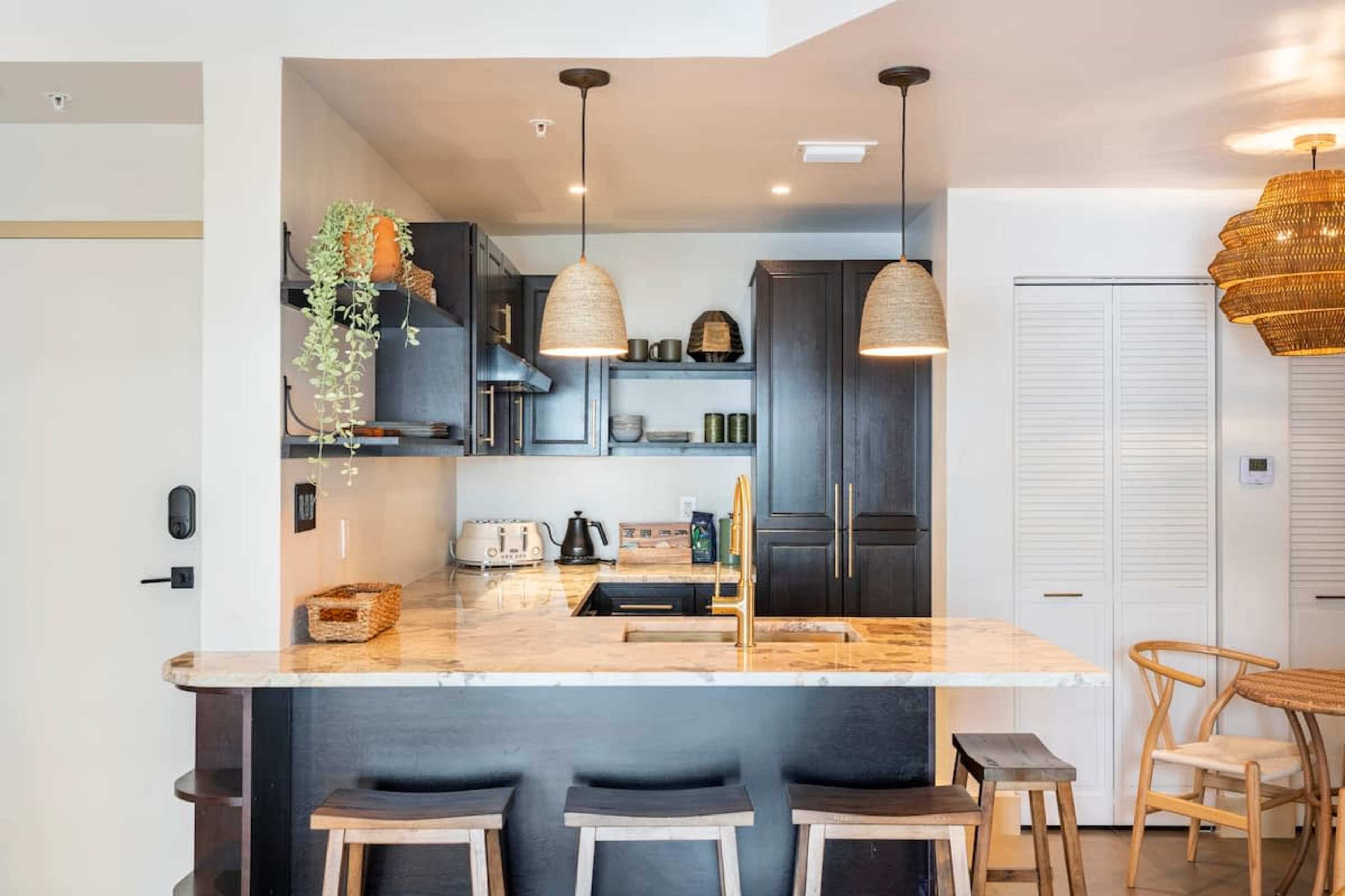 The image shows a modern kitchen with dark cabinetry, a marble countertop, and wooden stools at a bar.