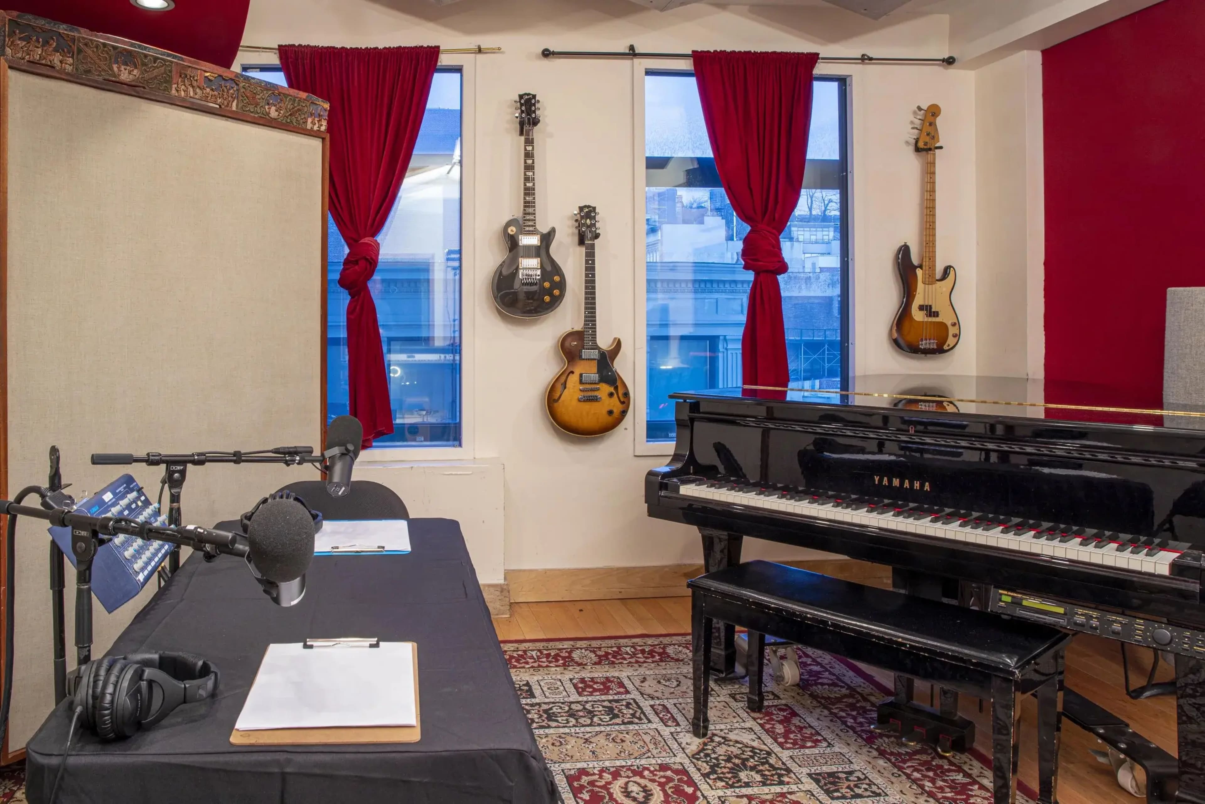 The image features a music studio with a black piano, guitars mounted on the wall, a microphone setup on a table, and large windows with red curtains.