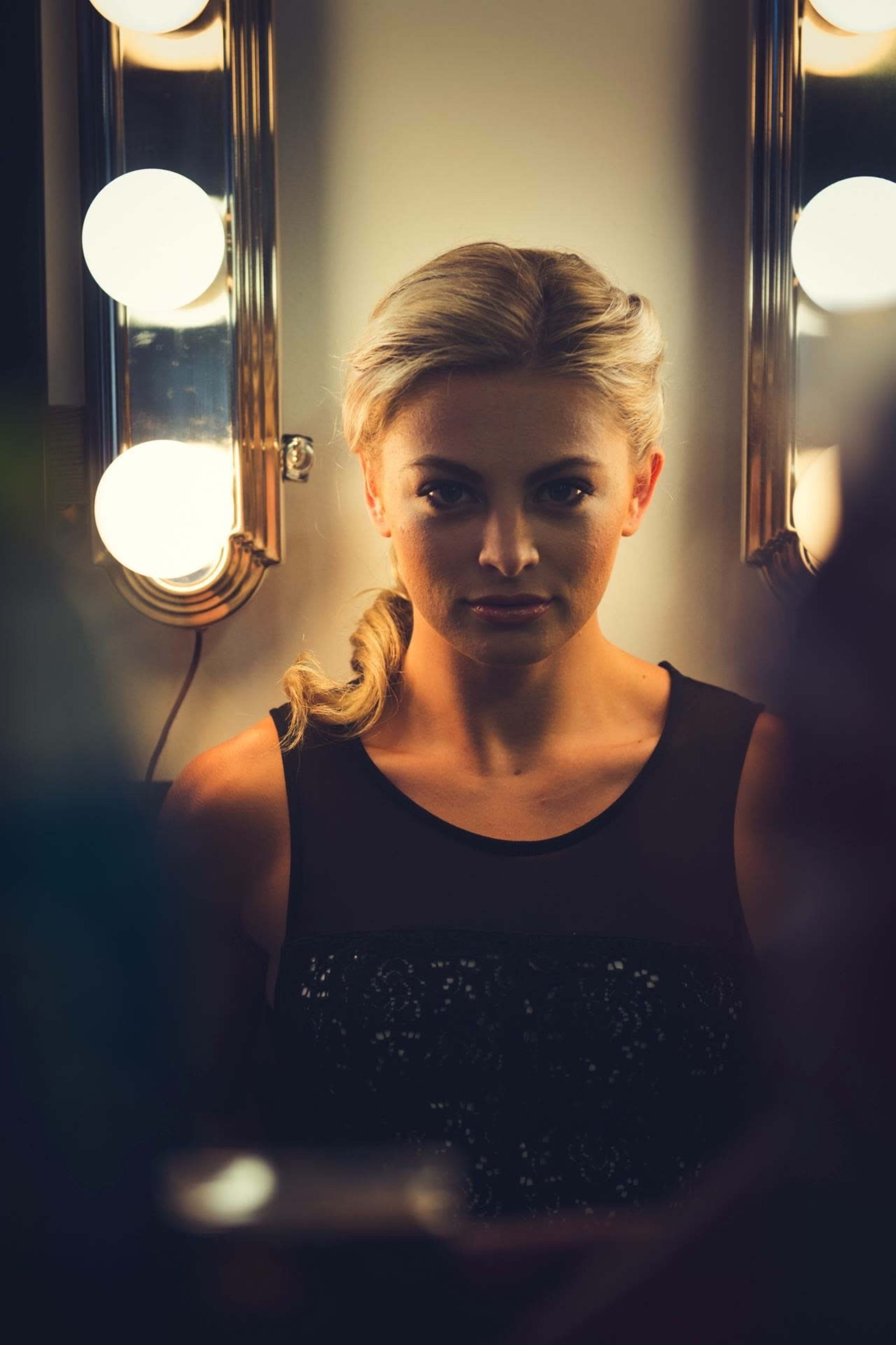 A woman with blonde hair is seated in front of a mirror surrounded by bright light bulbs.