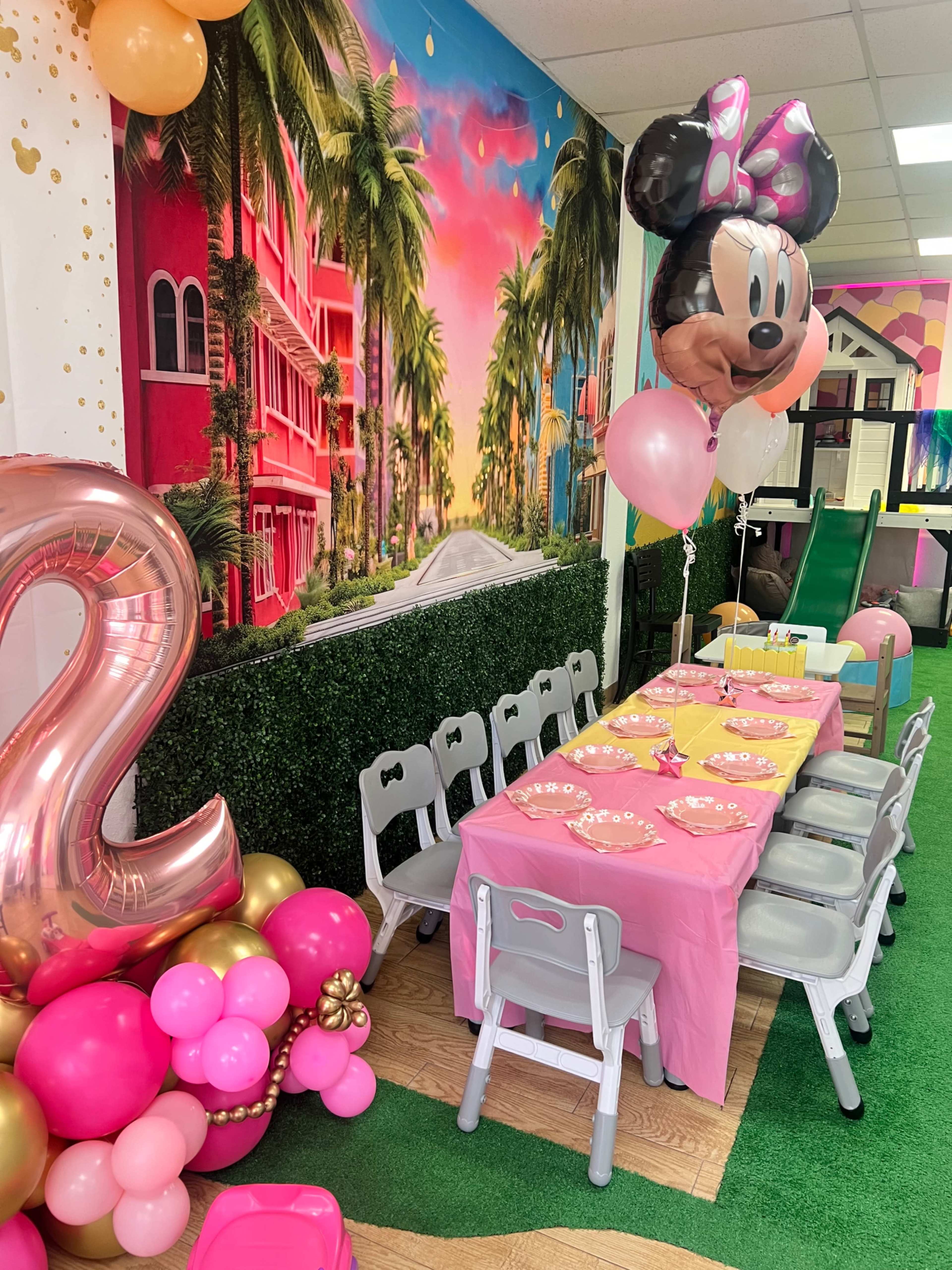 A colorful party setup features a decorated table with pink tablecloths, balloons, and a vibrant mural of palm trees and buildings in the background.
