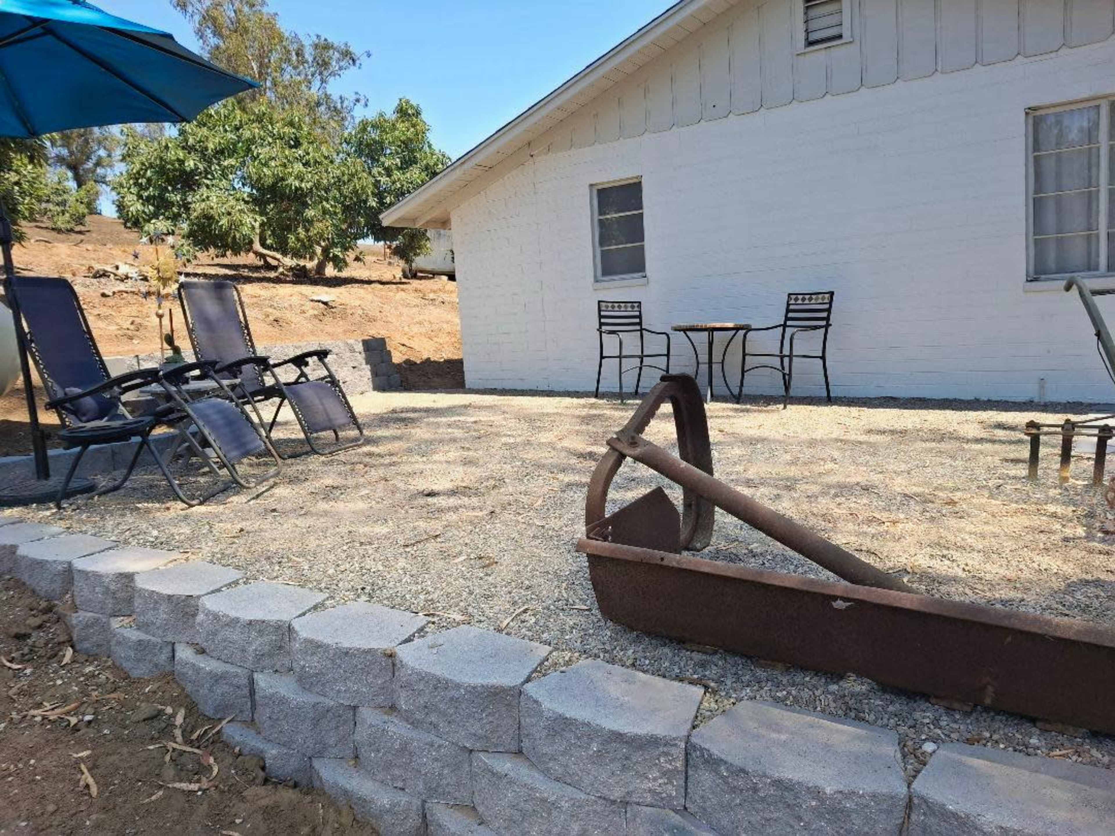 The image shows a gravel patio area in front of a white house, featuring two lounge chairs, a small table with chairs, and an old rusted piece of equipment.