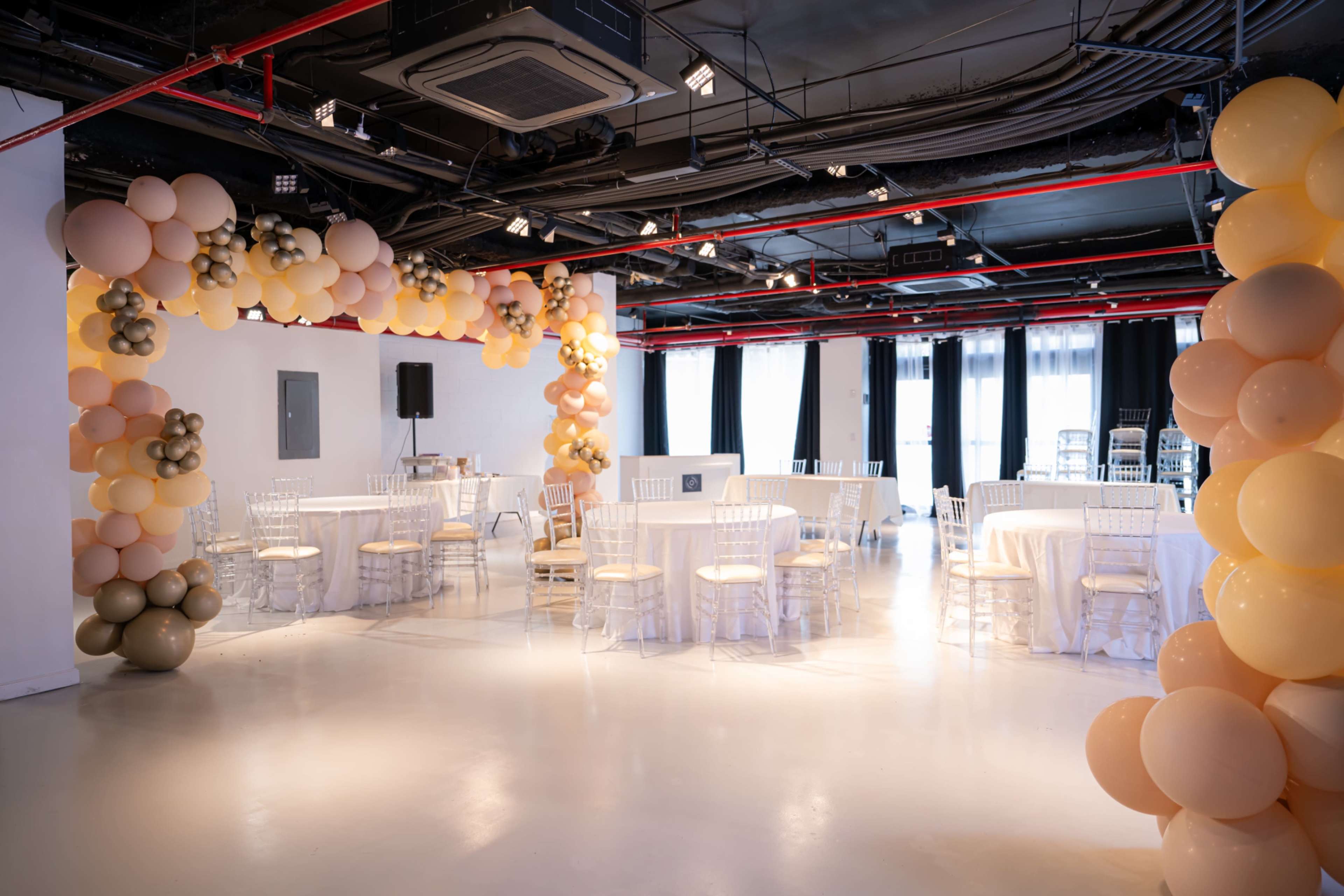 A spacious event hall features circular tables with white tablecloths surrounded by balloon decorations in varying colors.