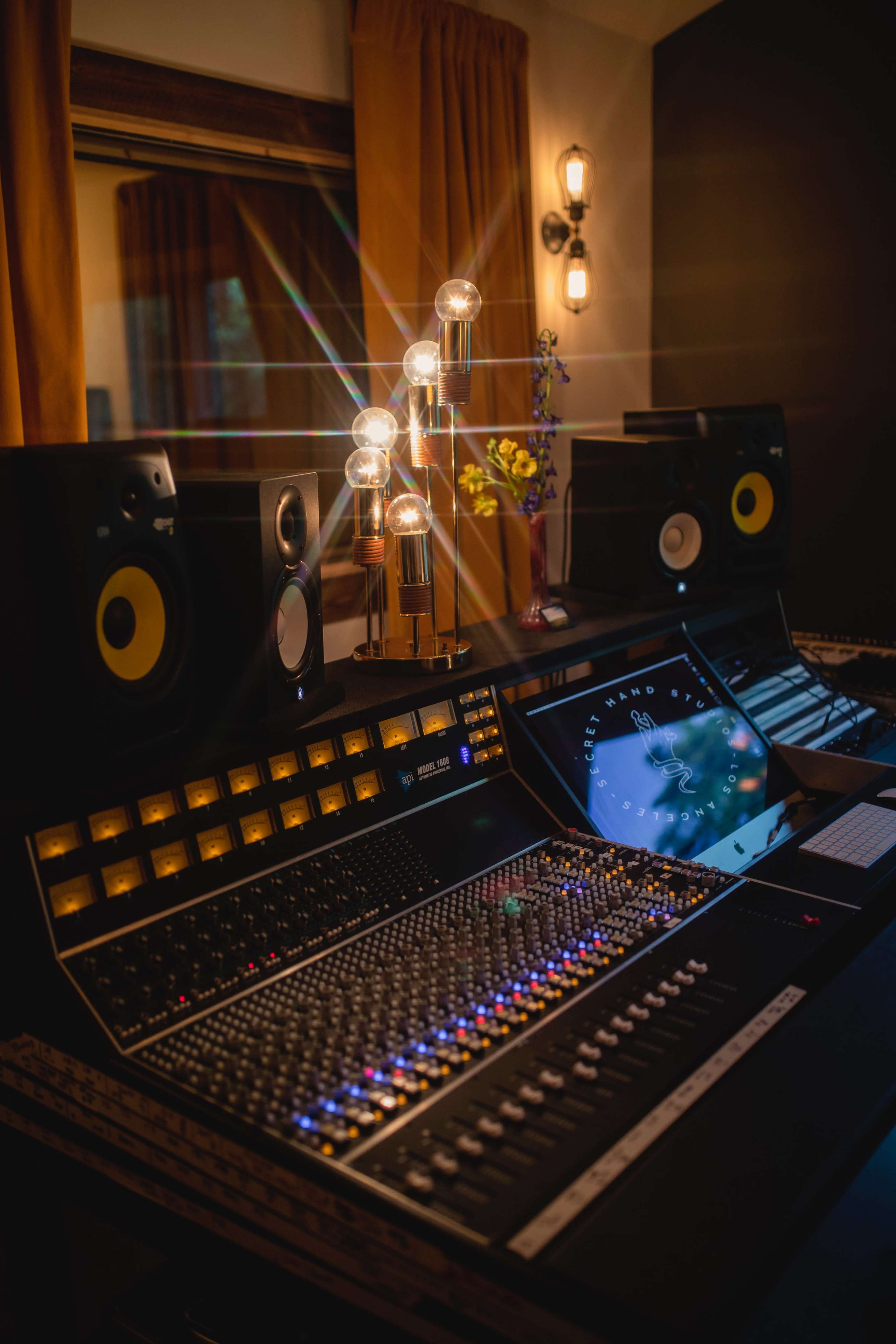 The image shows a modern music studio with a large mixing console, illuminated speakers, and a digital screen, set in a room with warm lighting and decorative curtains.