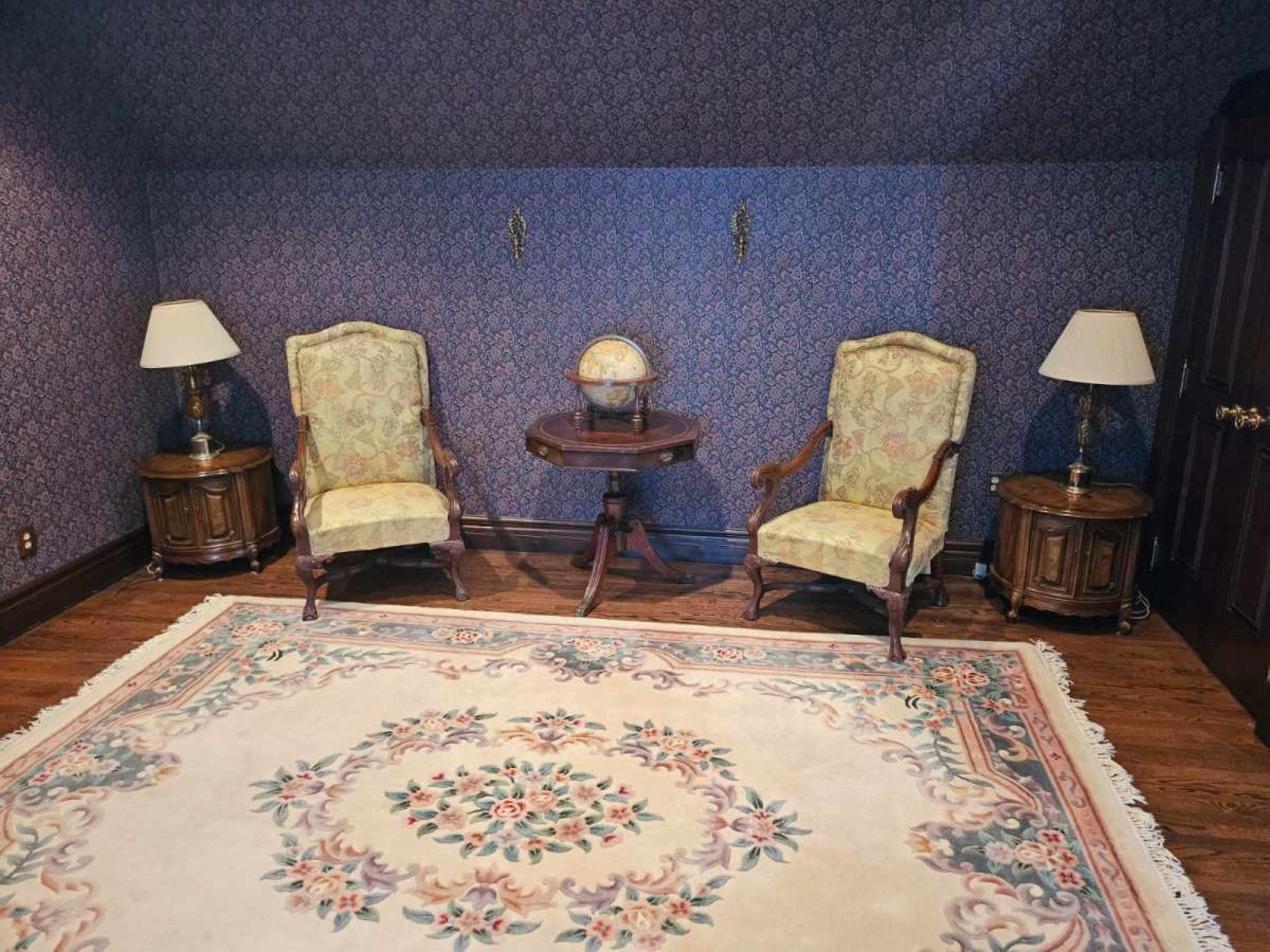 A room with two upholstered chairs, a small round table, a globe, two lamps, and a decorative rug on wooden flooring.