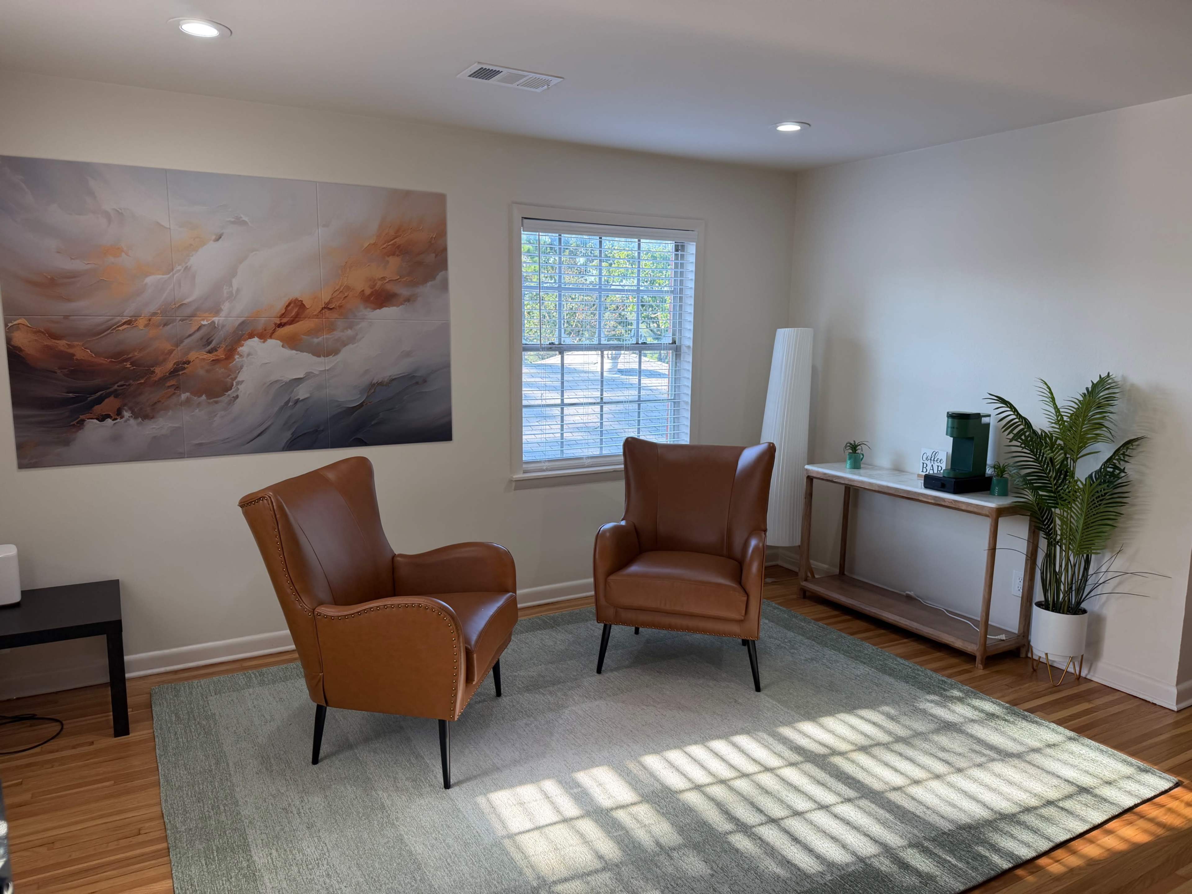 The image shows a bright room with two brown armchairs facing each other, a painting on the wall, a window letting in natural light, and a small wooden table with a plant and decor.
