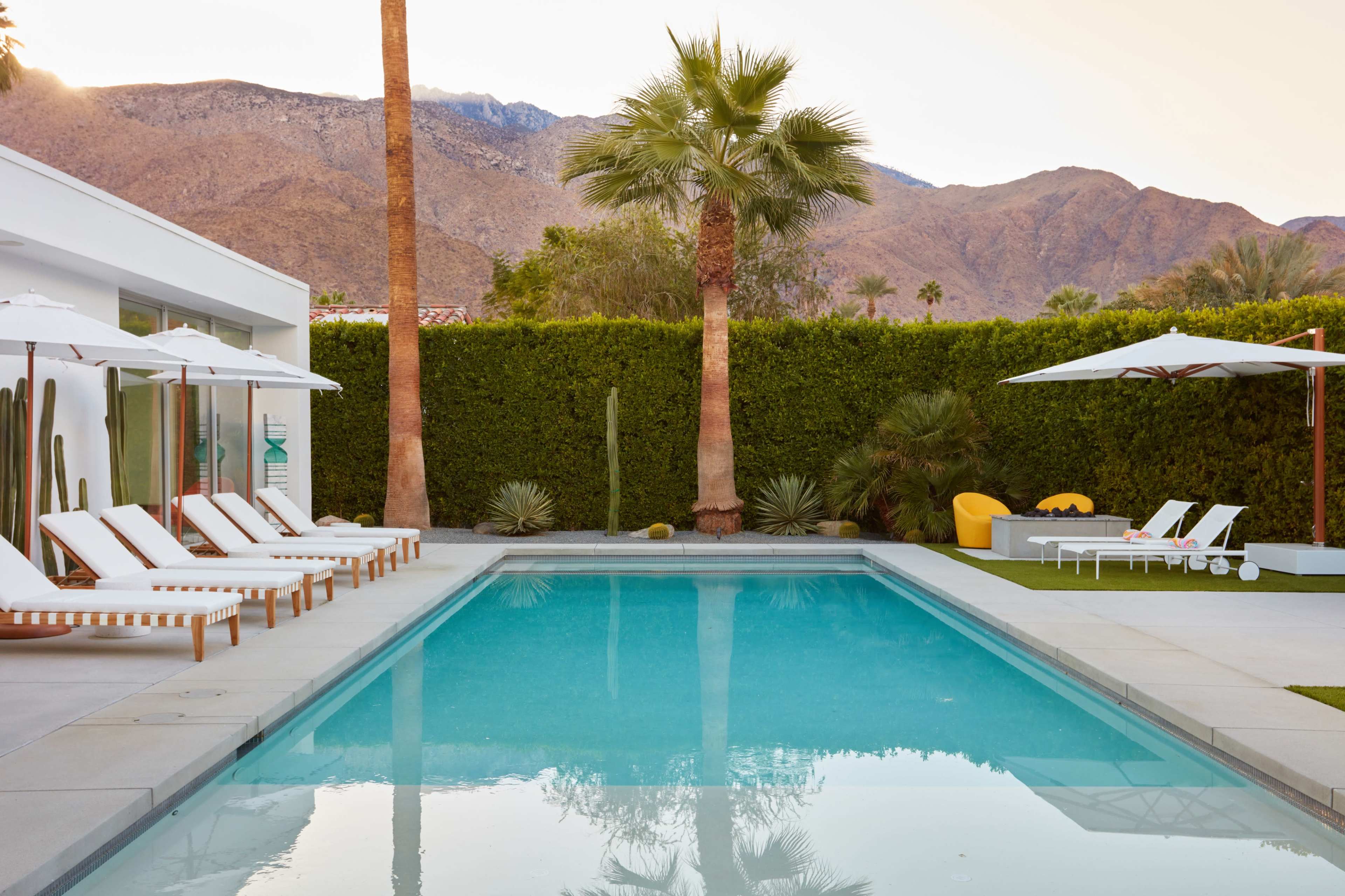 The image features a modern outdoor pool surrounded by lounge chairs and palm trees, with mountains in the background.