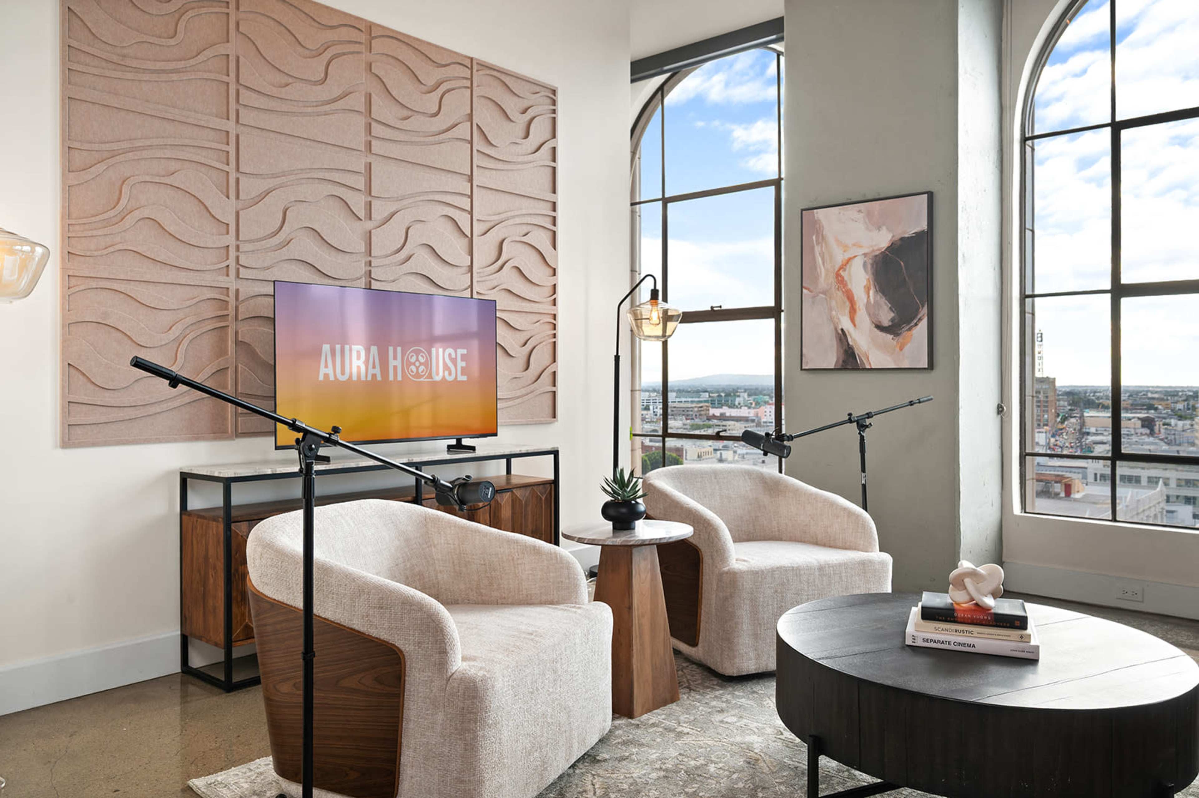 A living room features two modern chairs facing a television mounted on the wall, with a textured panel behind it and large windows providing a city view.