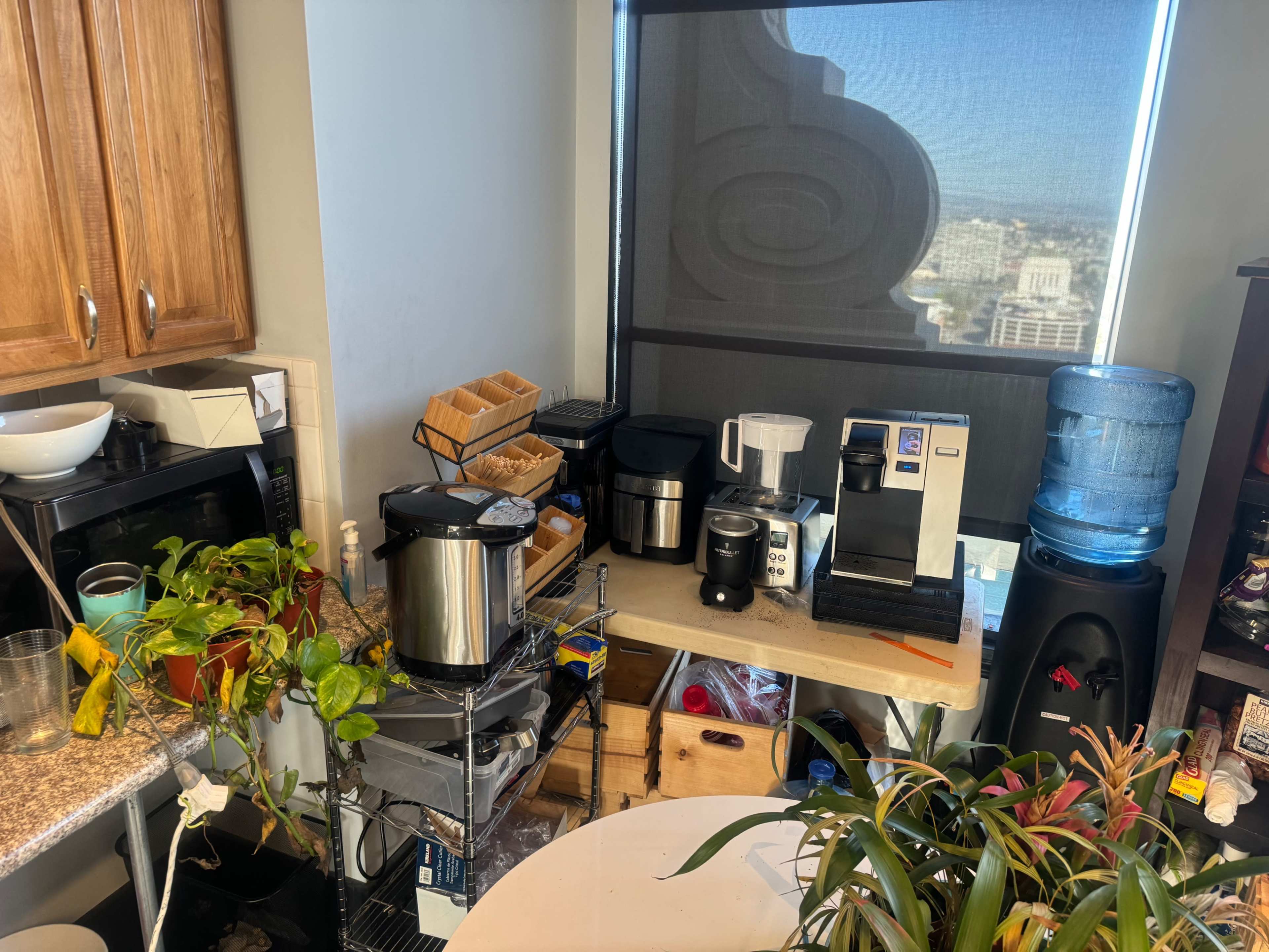 A cluttered kitchen corner features various coffee appliances, a water cooler, and plants near a window with a city view.
