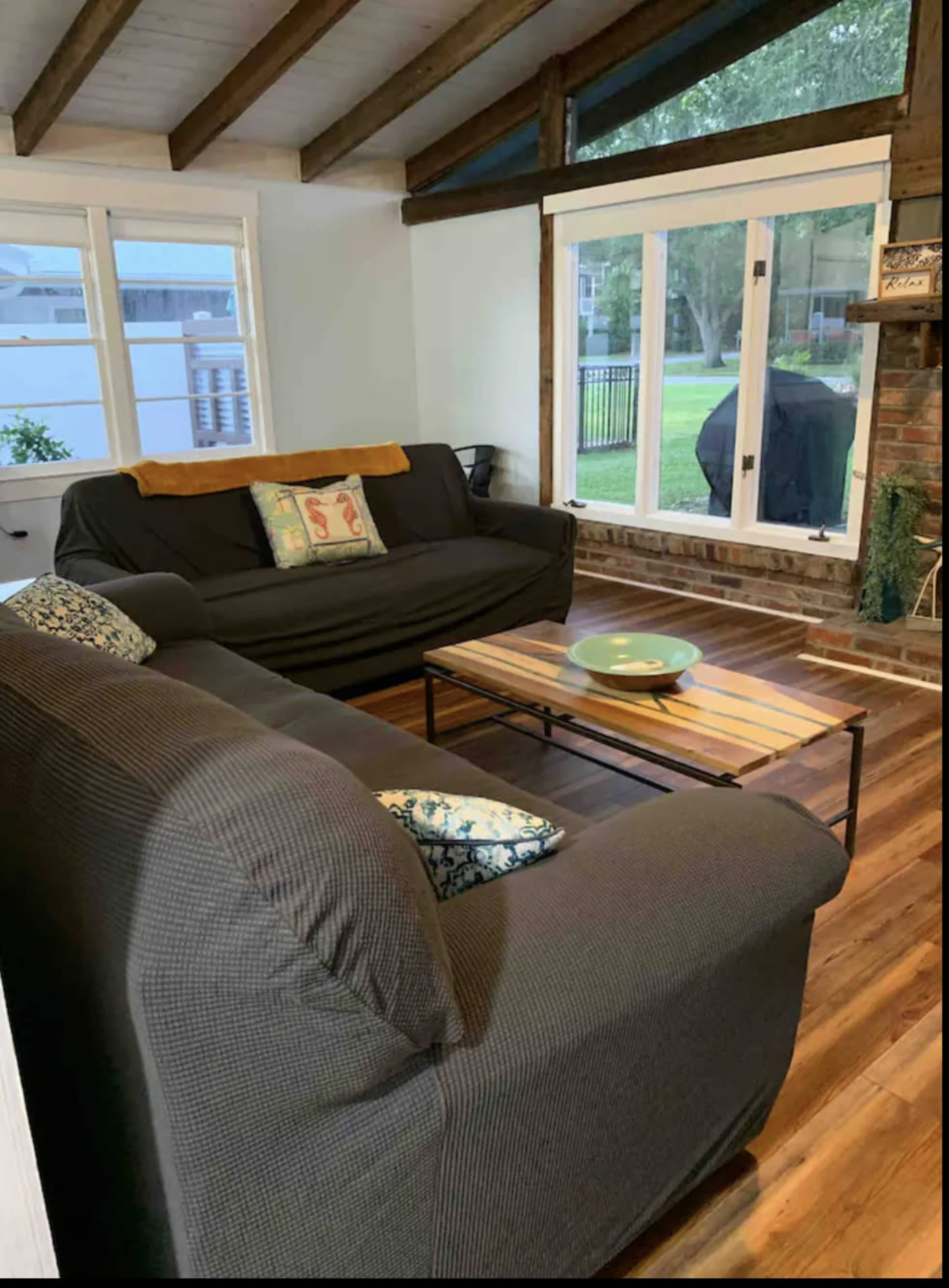 The image shows a cozy living room with two dark sofas facing a coffee table, wooden beams on the ceiling, and large windows looking out to a patio.