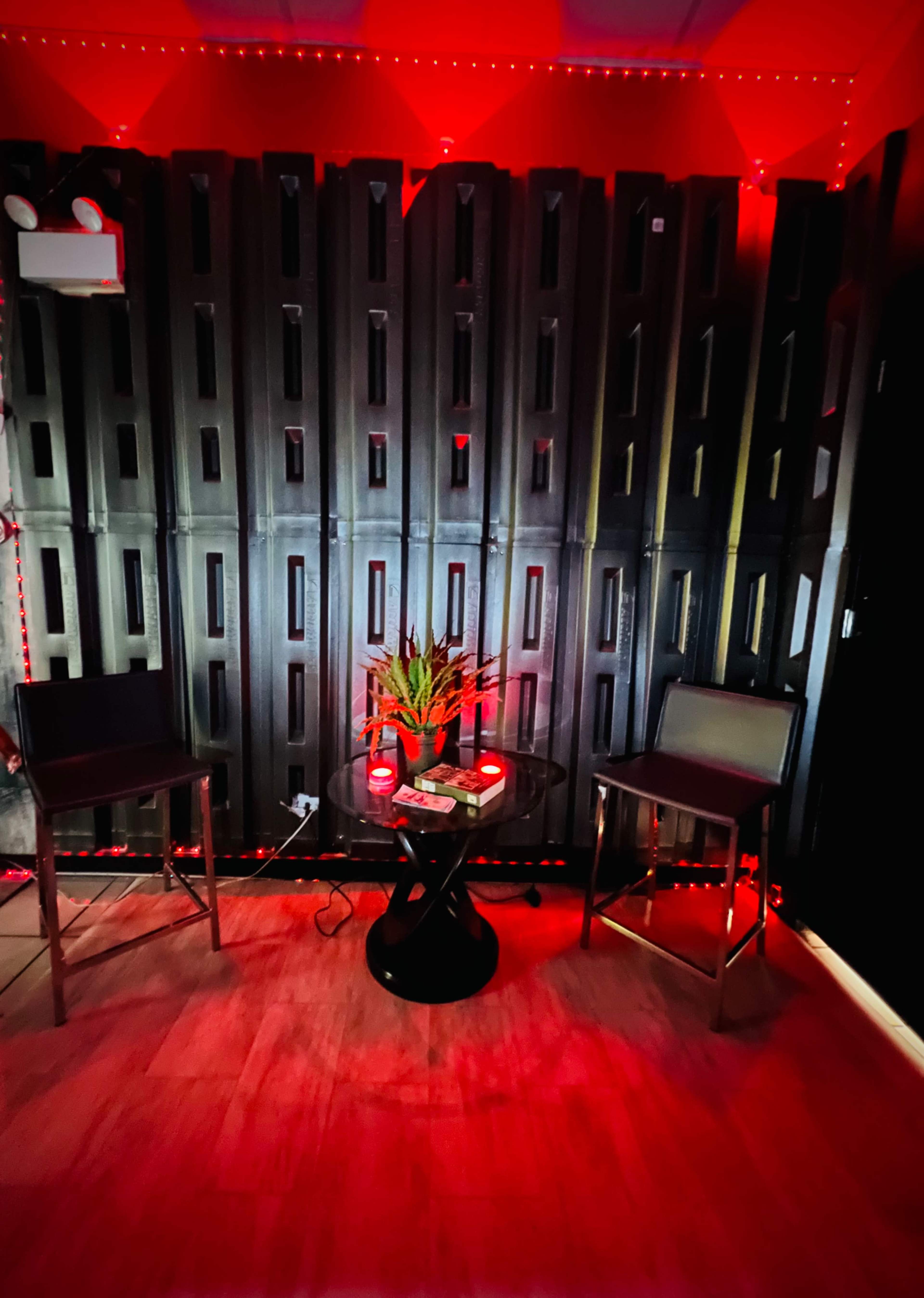 The image depicts a dimly lit room featuring two bar stools on either side of a small round table adorned with decorative items, all set against a backdrop of vertical black panels illuminated by red lights.