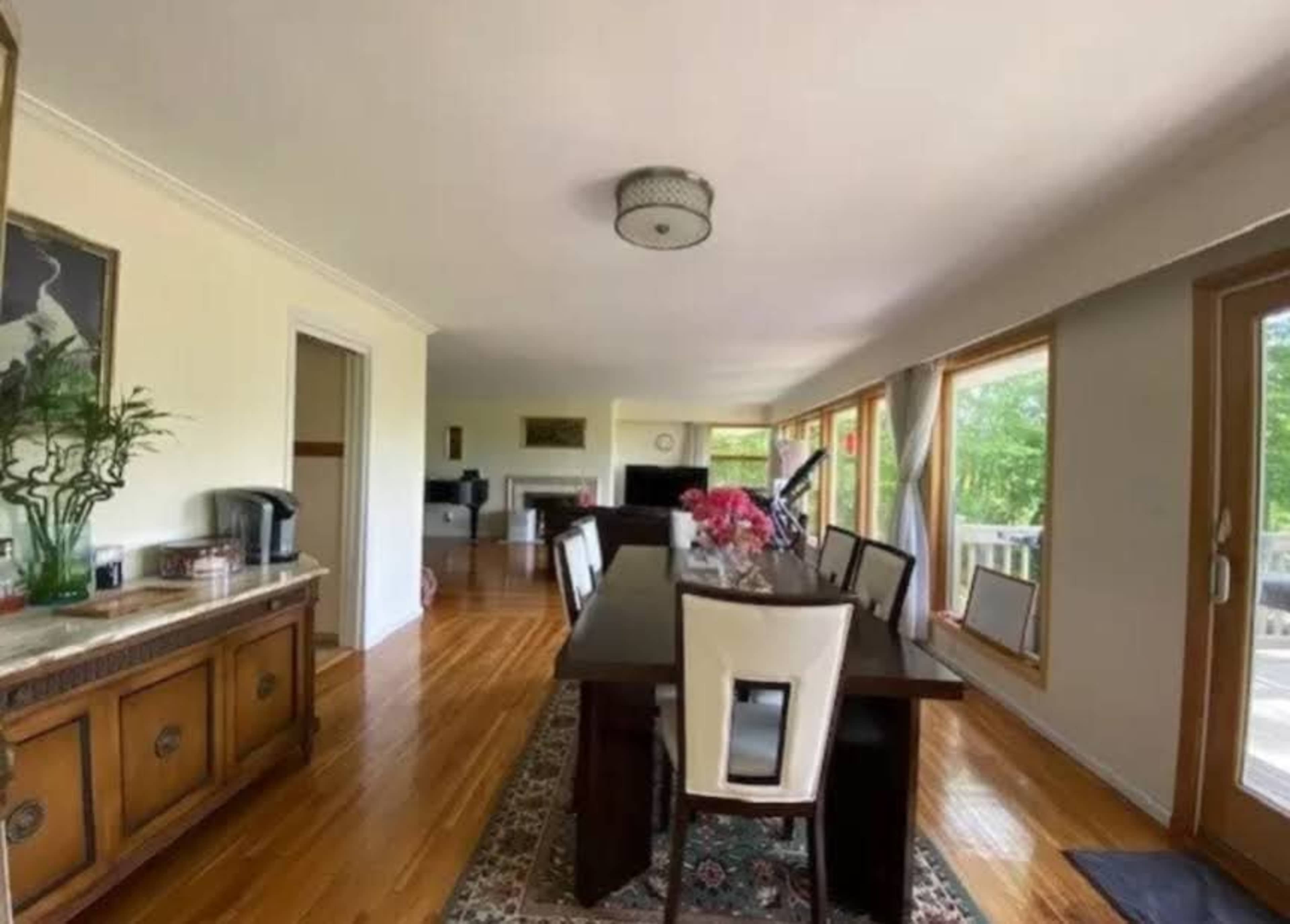 The image shows a spacious dining room with a long wooden table, surrounded by chairs, and large windows that let in natural light.