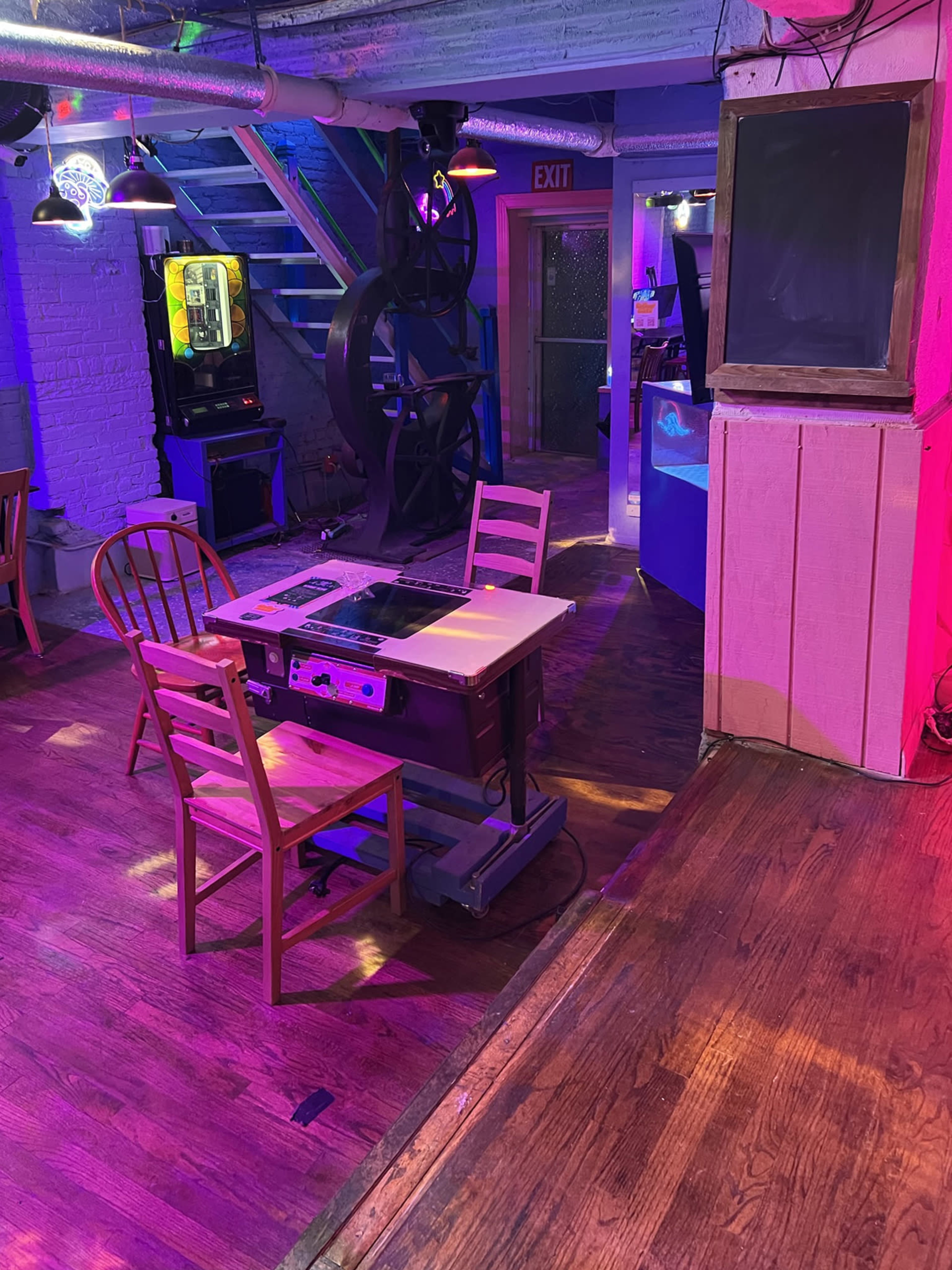 The scene depicts a dimly lit room featuring a vintage table and two wooden chairs near a spiral staircase, with colorful lighting and various retro decor elements.