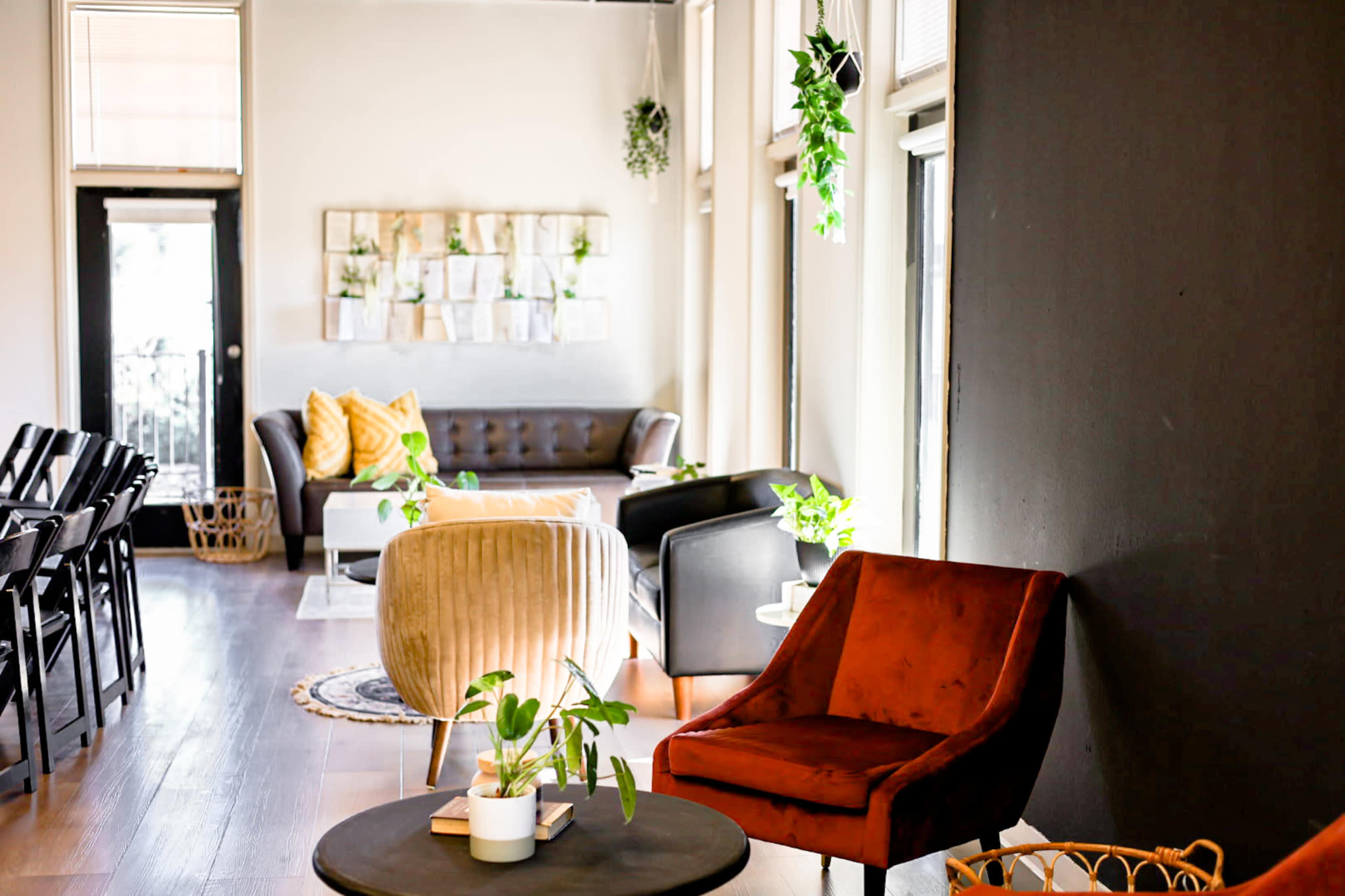 The image shows a modern, well-lit lounge area featuring various seating arrangements, plants, and a decorative wall with framed items.