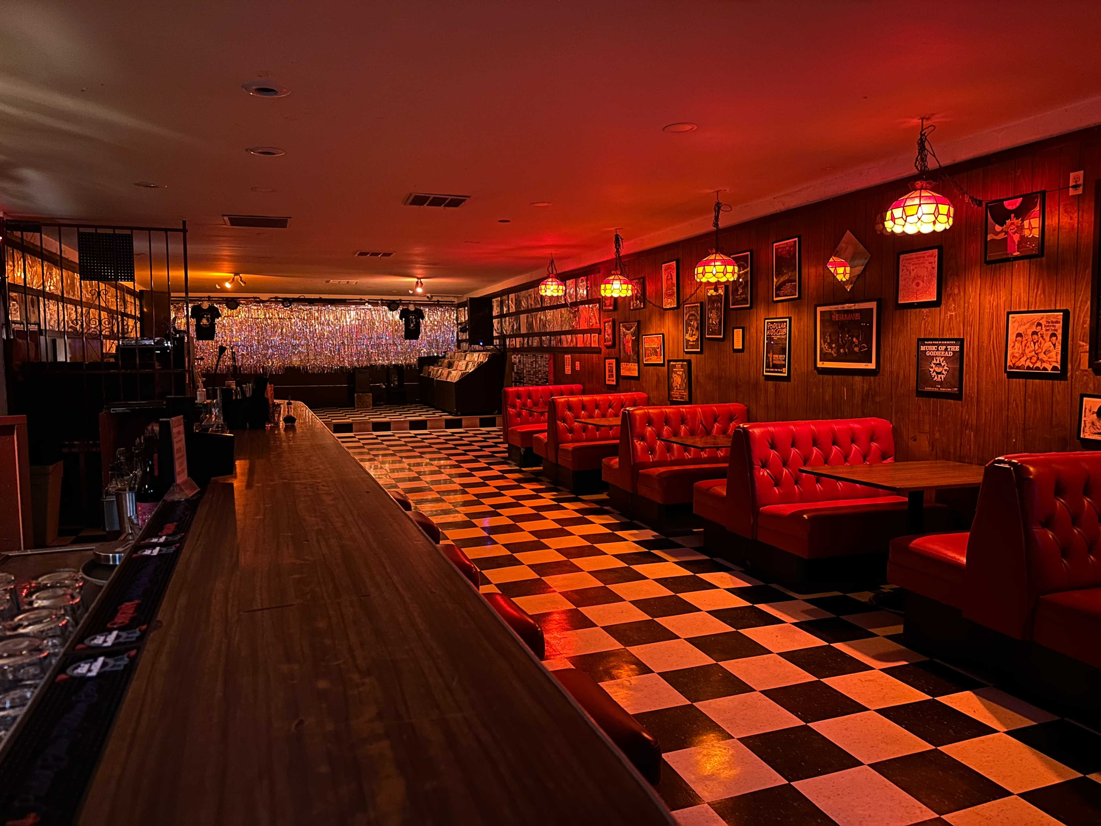 The image shows a dimly lit bar with red booths, a long wooden counter, and black-and-white checkered flooring, decorated with framed artwork on the walls.