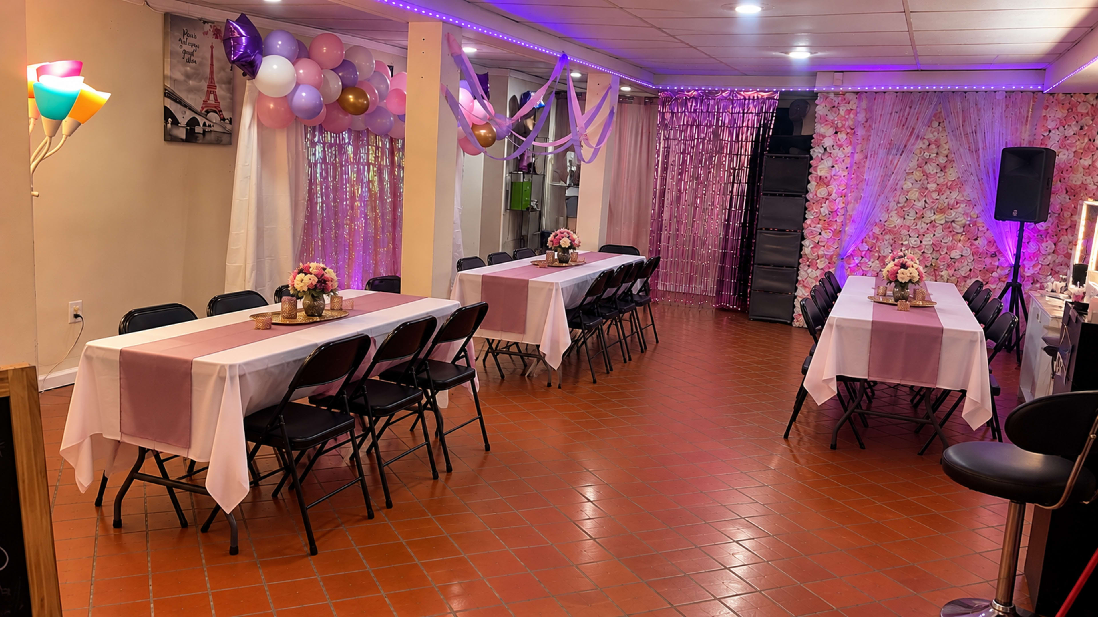 The room features decorated tables with white and pink tablecloths, surrounded by balloons and floral arrangements, set up for a celebration.