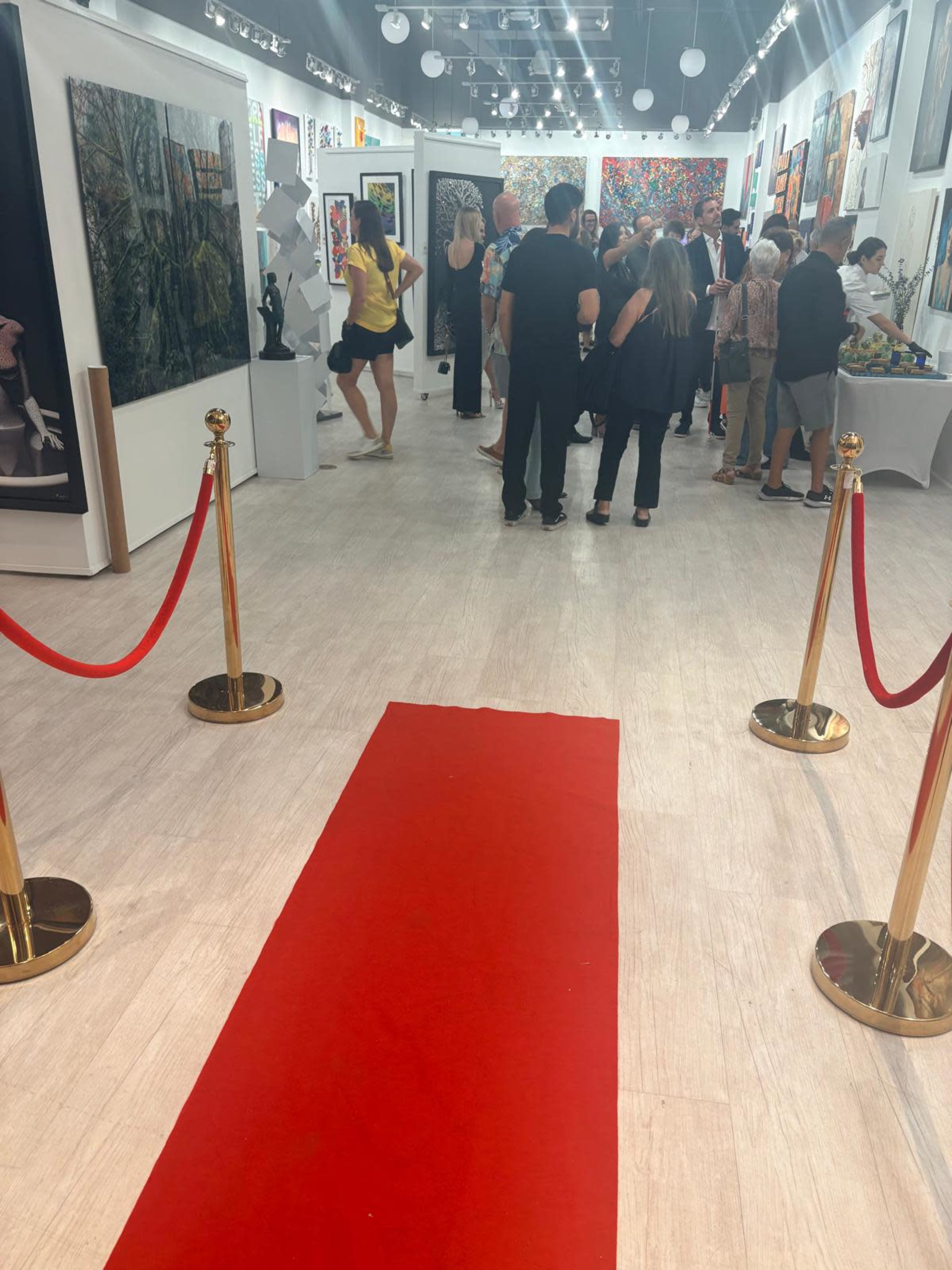 The image shows a brightly lit art gallery with people mingling among various artworks, framed pieces displayed on the walls, and a red carpet leading into the space.