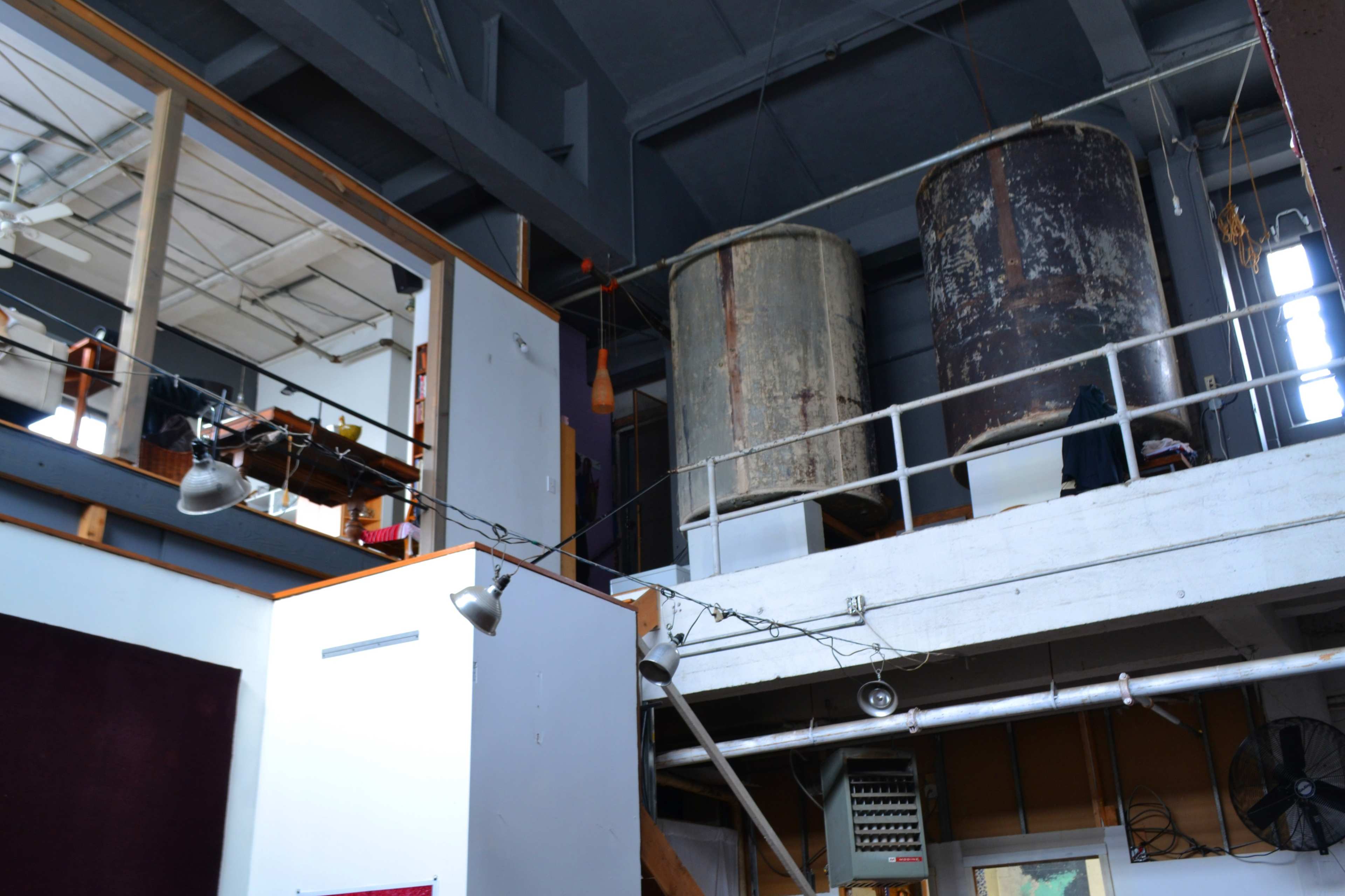 A loft-like space with a balcony overlooking two large, rusty storage tanks and exposed ducts.