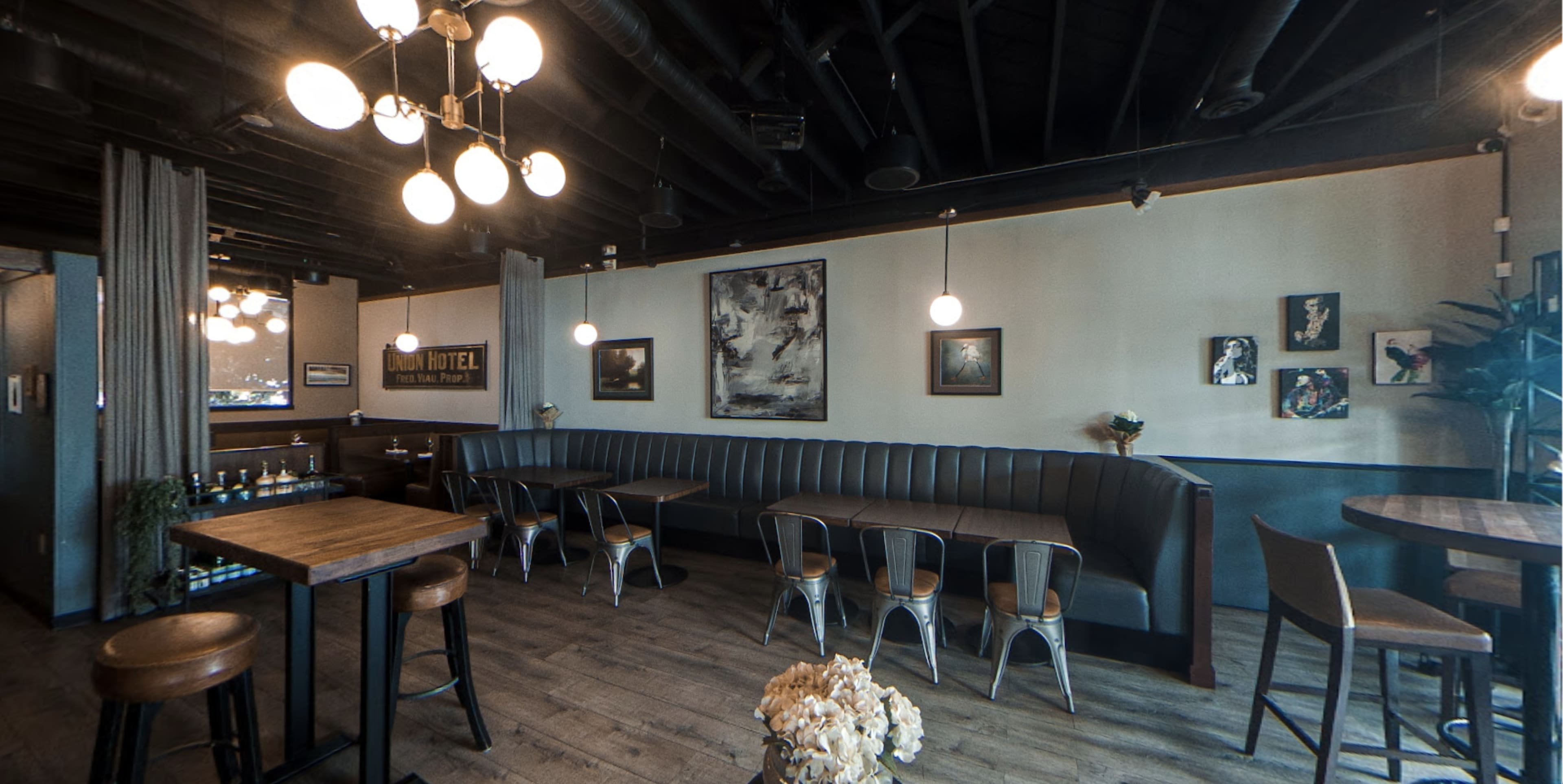 A modern café interior with a curved leather booth, wooden tables, and various artwork on the walls, illuminated by hanging light fixtures.