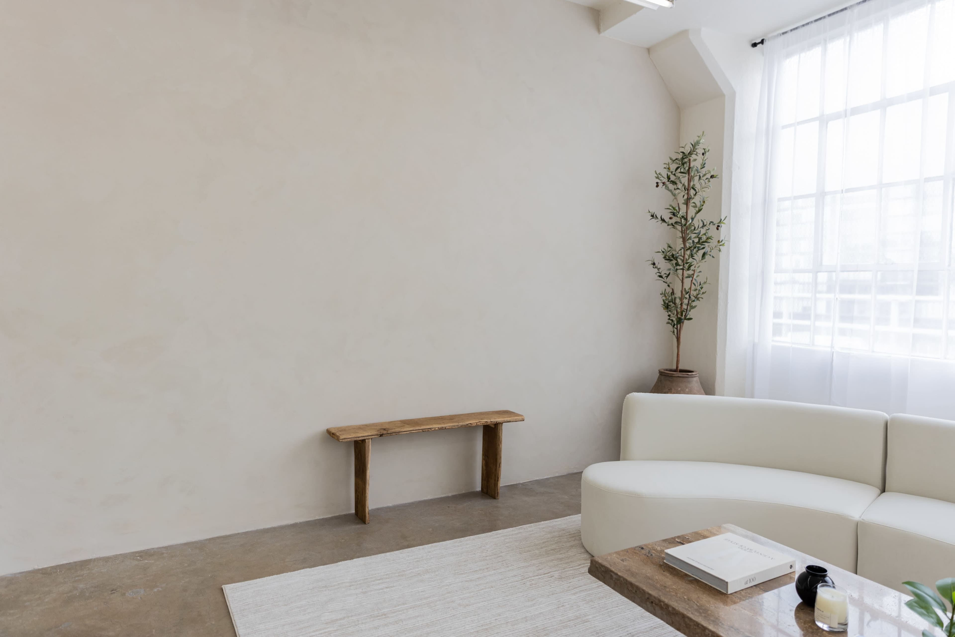 A minimalist room with a light-colored wall, a wooden bench, a white sofa, and a potted plant near a large window.
