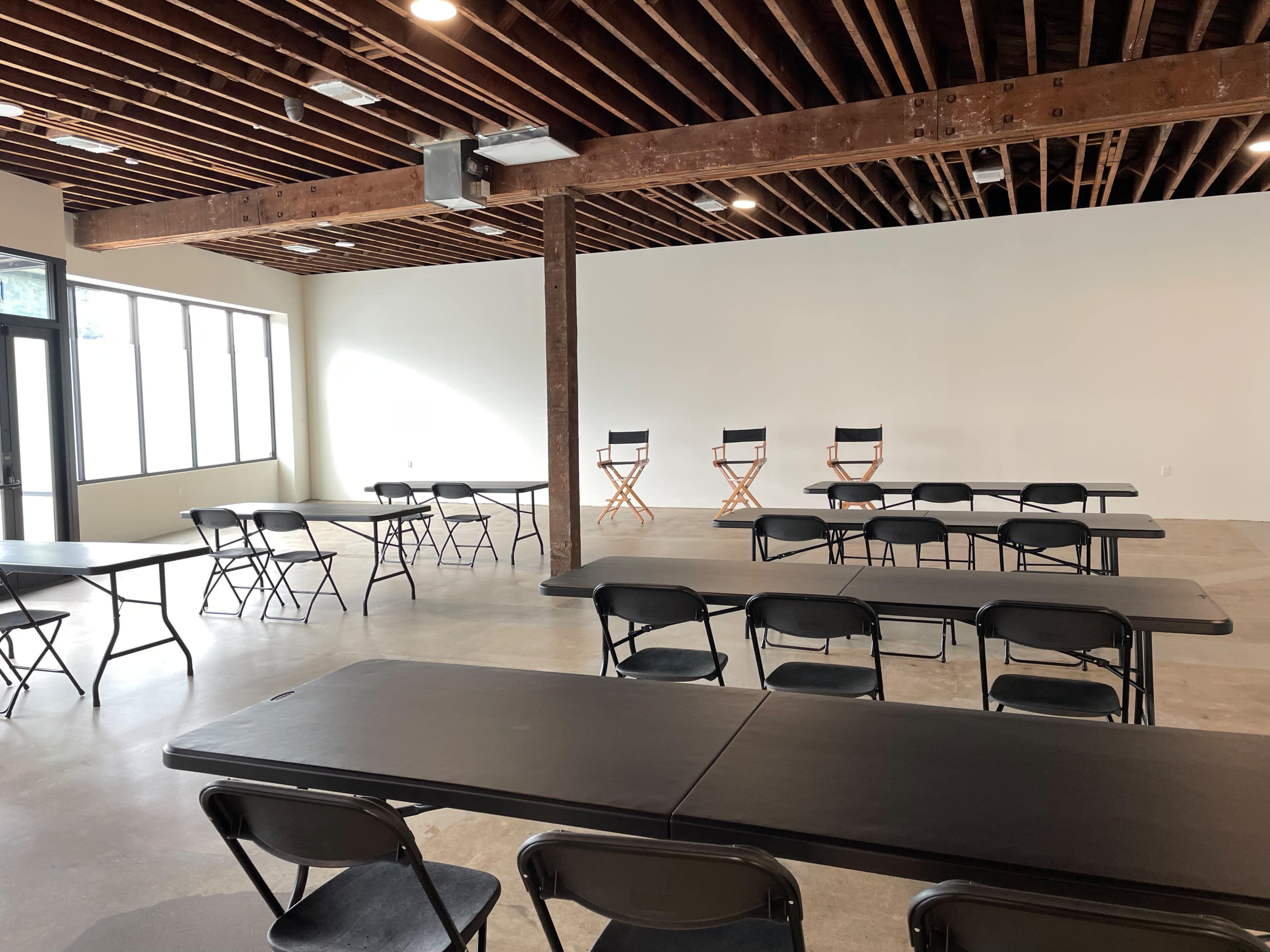 A spacious room with bare walls, exposed wooden beams, and several black folding tables and chairs arranged facing a blank wall where three director's chairs are positioned.