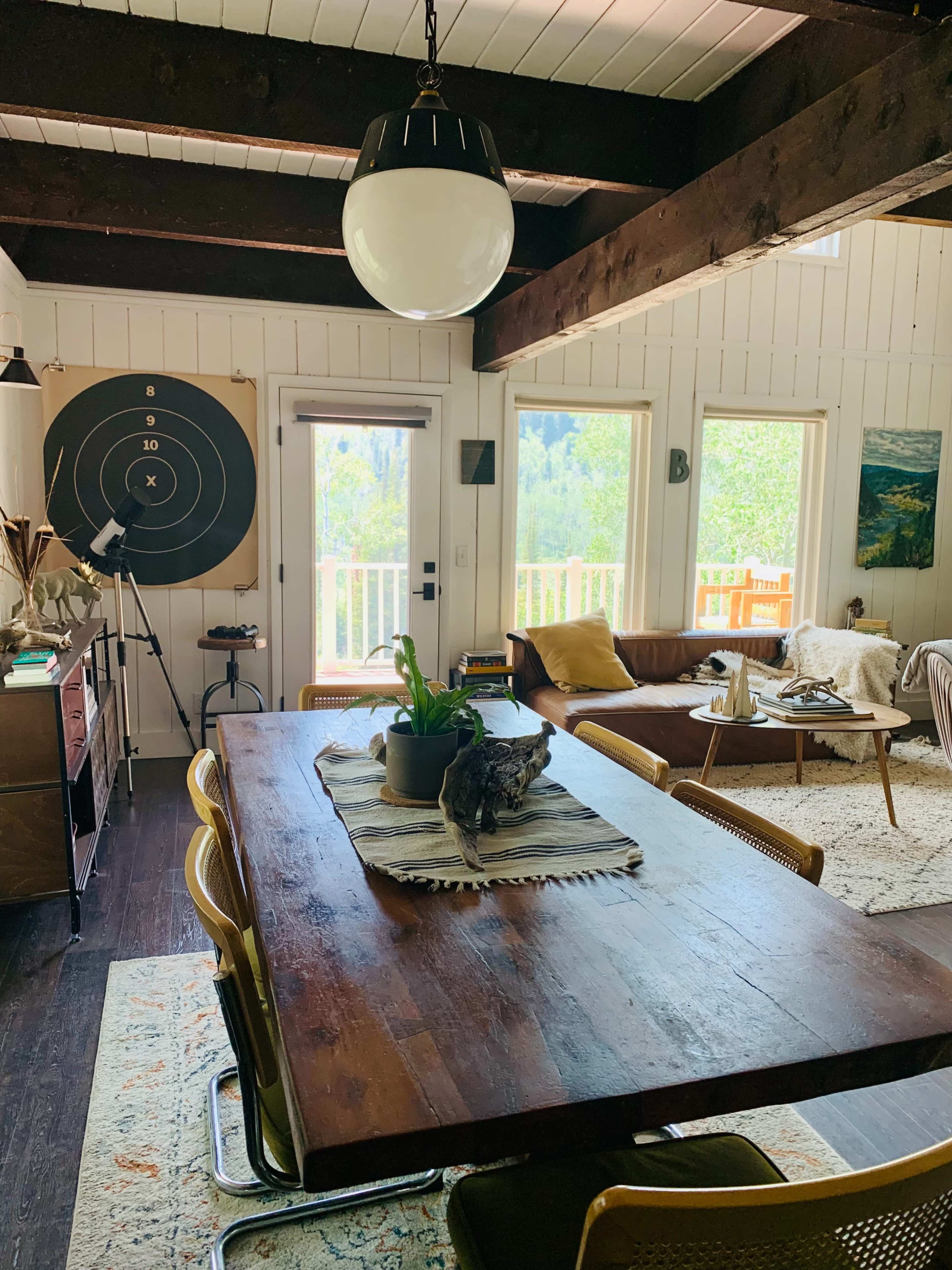 The image shows a cozy interior of a room featuring a wooden dining table, a leather sofa, and large windows with views of greenery outside.