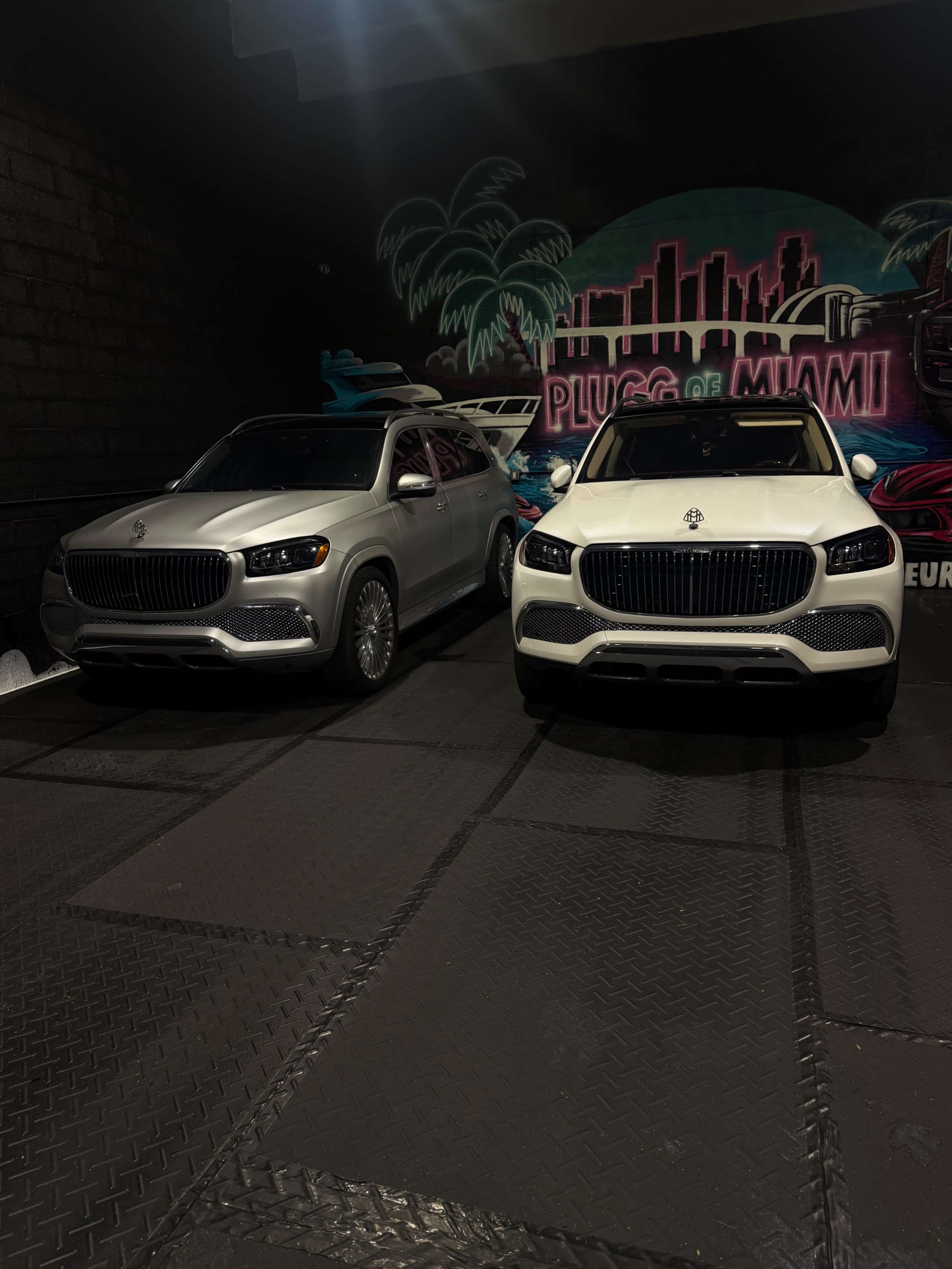 Two luxury SUVs are parked side by side in a dimly lit garage with a mural of Miami in the background.