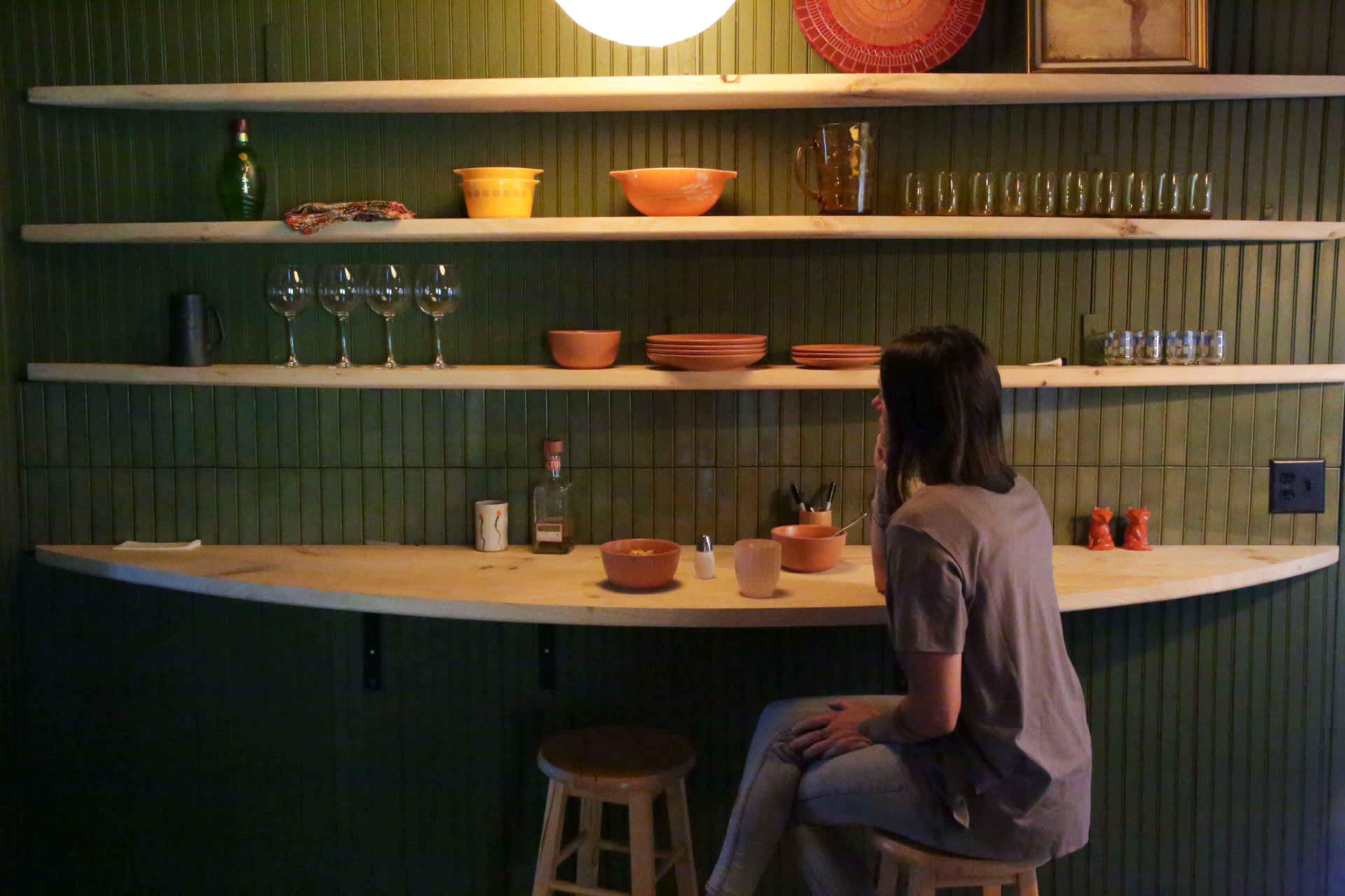 A woman sits at a curved wooden countertop in a dimly lit room with shelves displaying dinnerware and glassware.