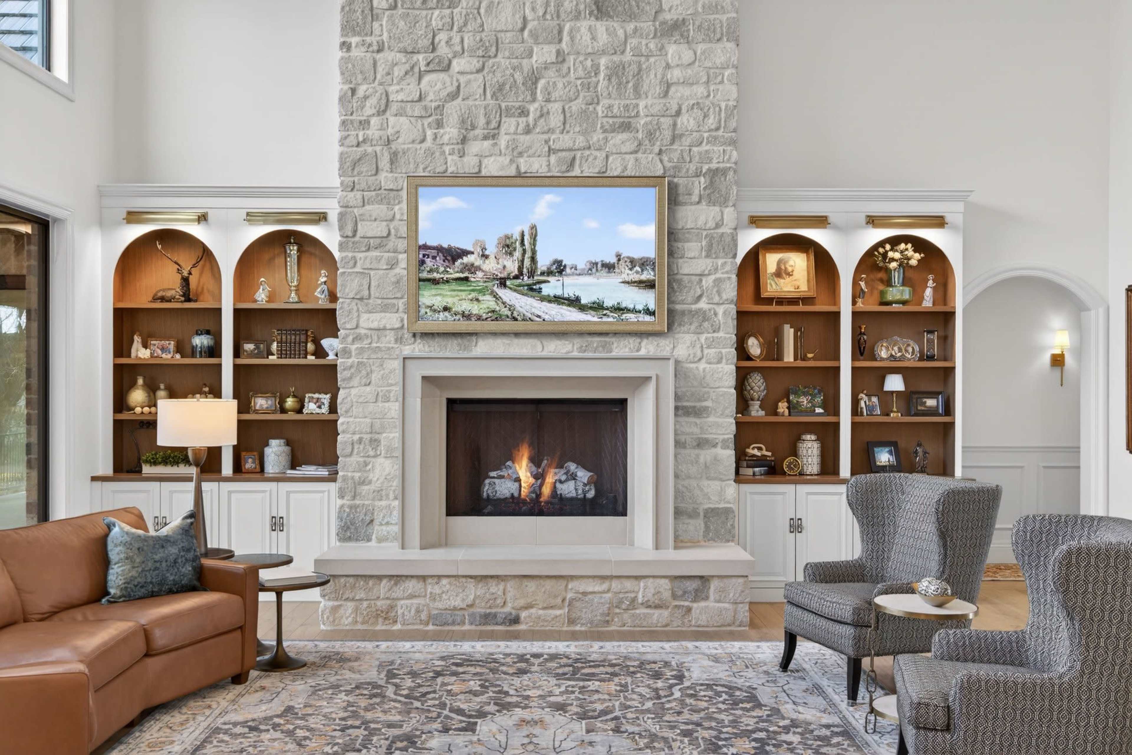 The image depicts a modern living room featuring a stone fireplace with a television above it, surrounded by built-in bookshelves displaying decorative items, a brown leather couch, and two patterned armchairs on a large area rug.