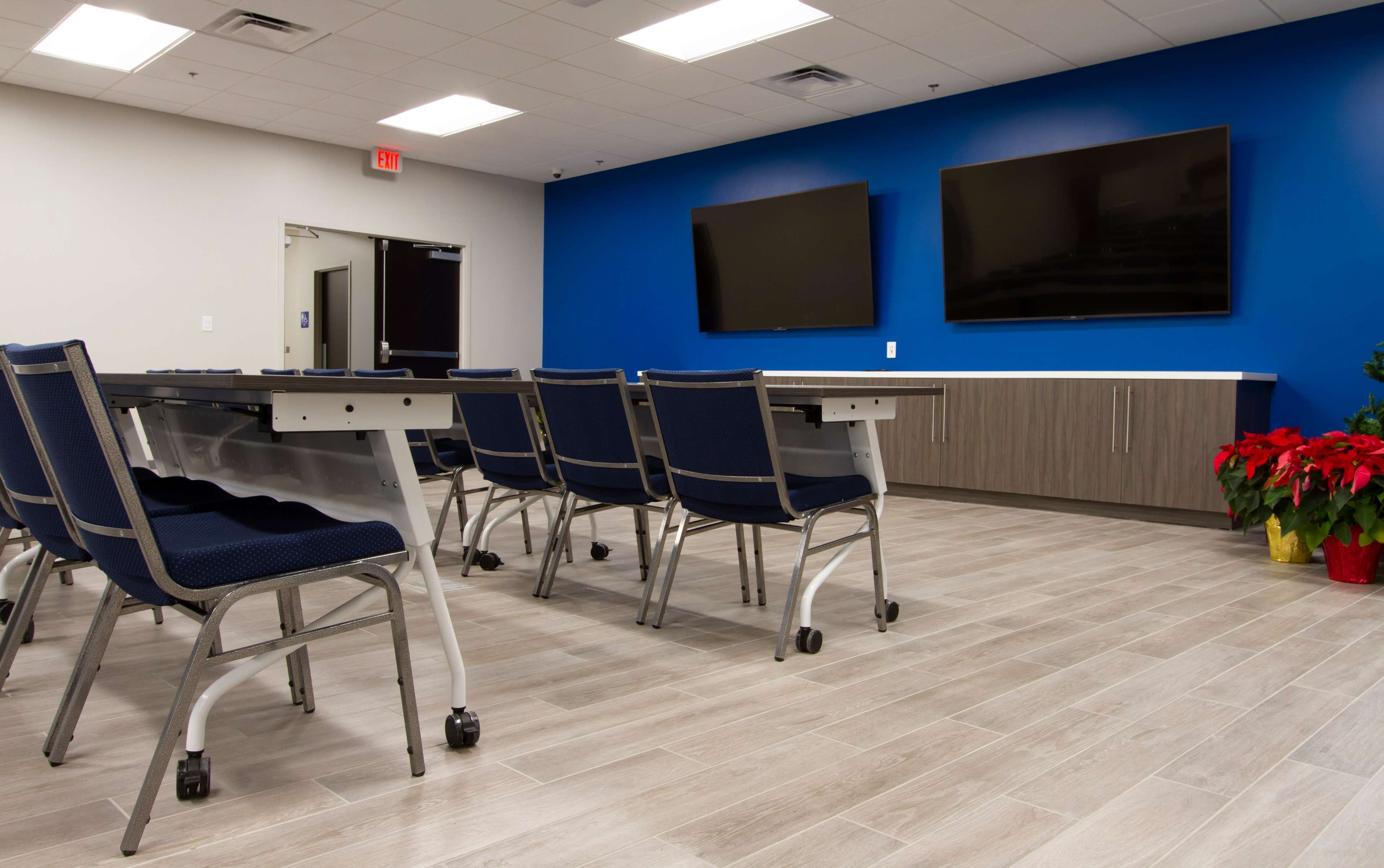 A modern meeting room with rows of chairs facing two wall-mounted televisions and a cabinetry unit, all set against a blue accent wall.