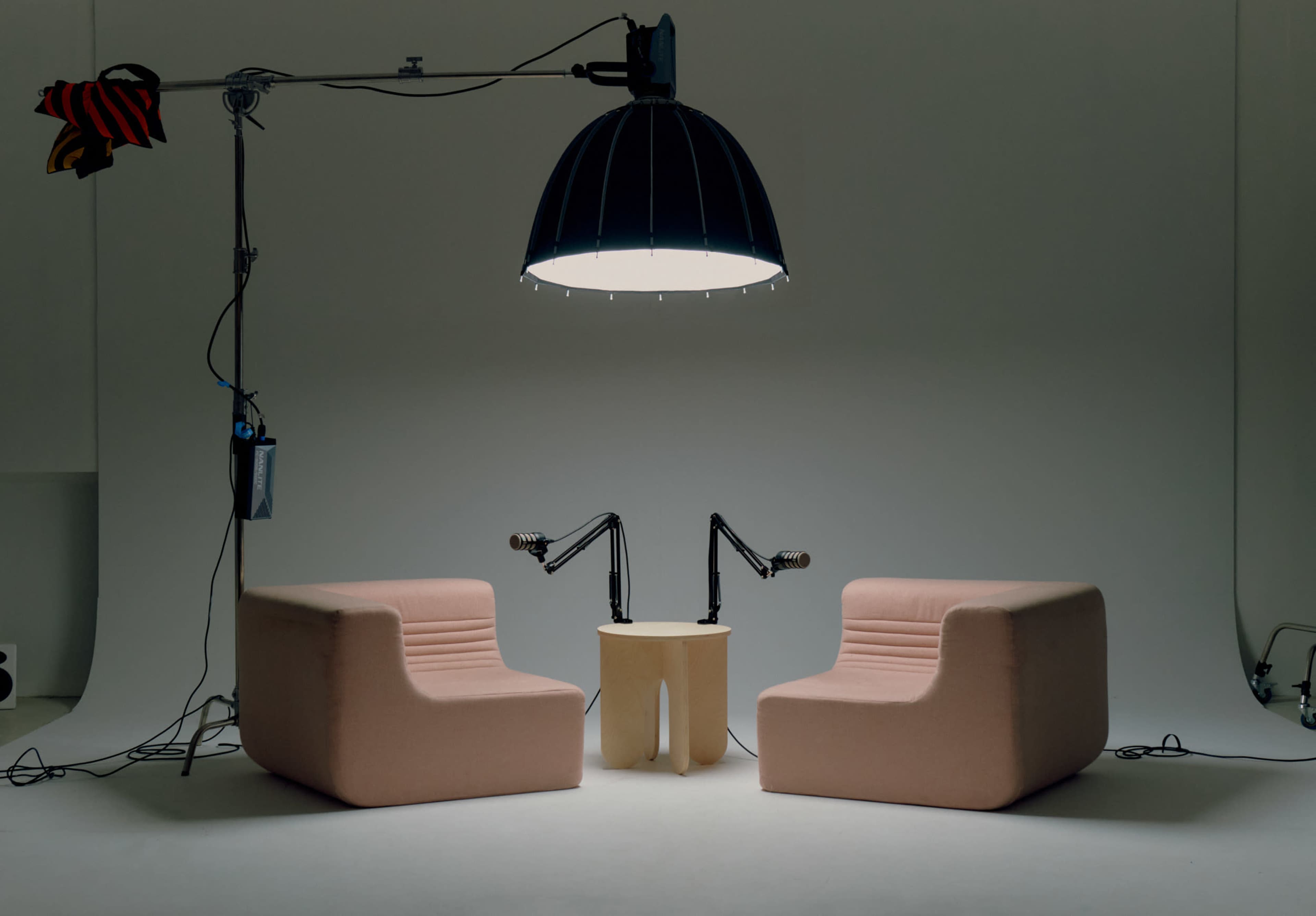 The image shows two pink, upholstered chairs facing each other, with a round wooden table between them, illuminated by a large pendant light in a minimalistic studio setting.