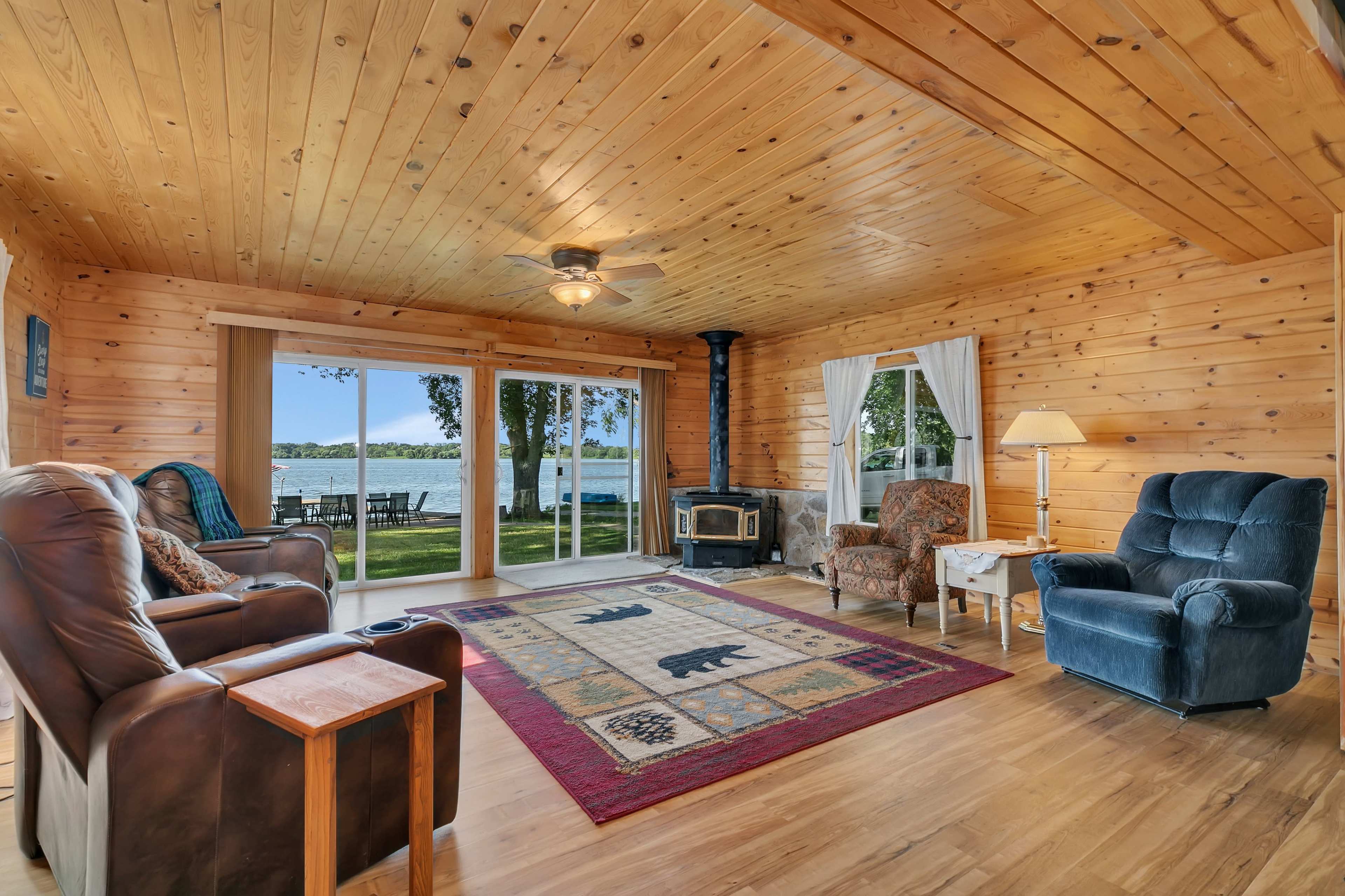 The room features wooden walls and ceiling, a fireplace, and large windows overlooking a lake.