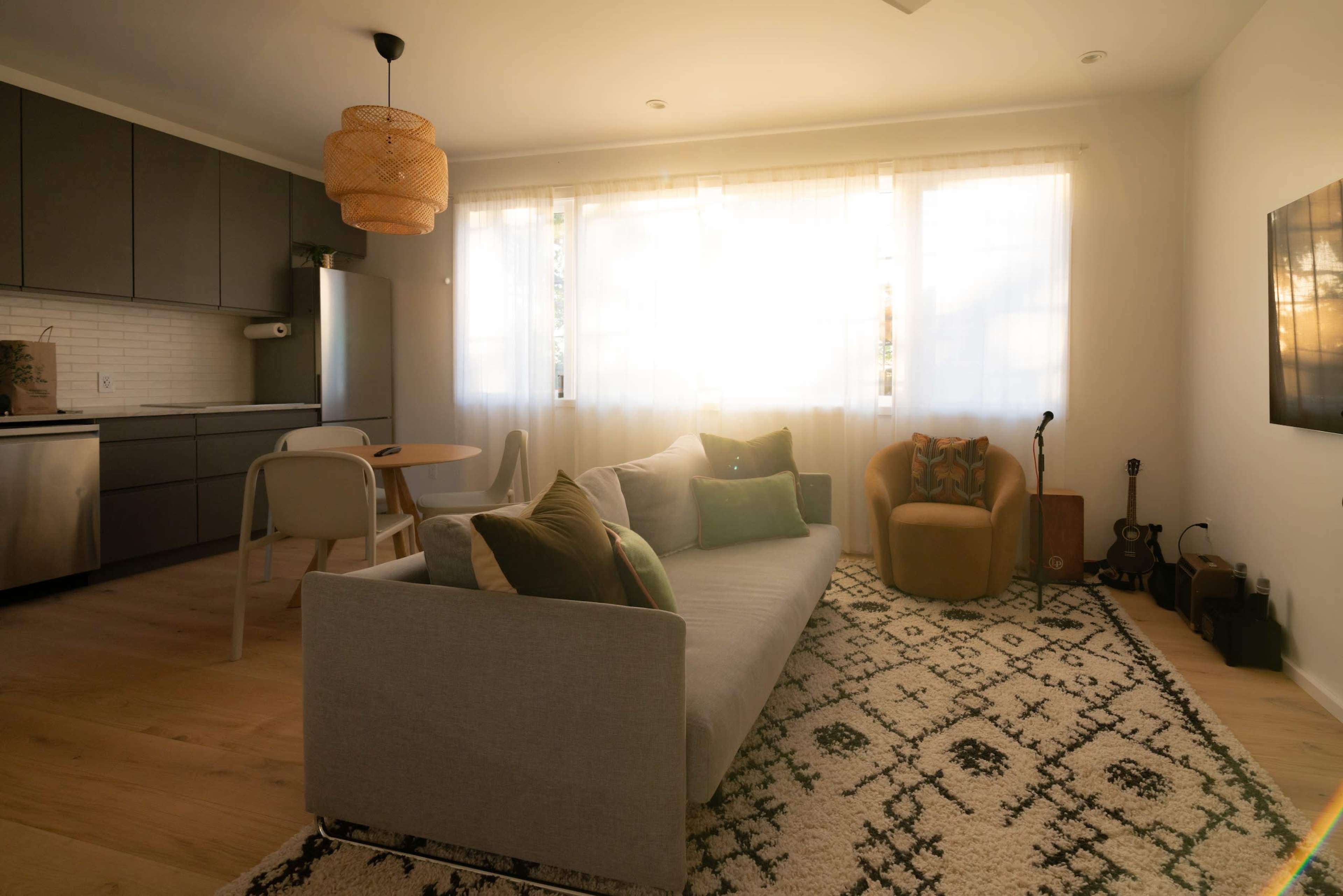 A bright living room with a gray sofa, a round dining table, and large windows allowing sunlight to illuminate the space.