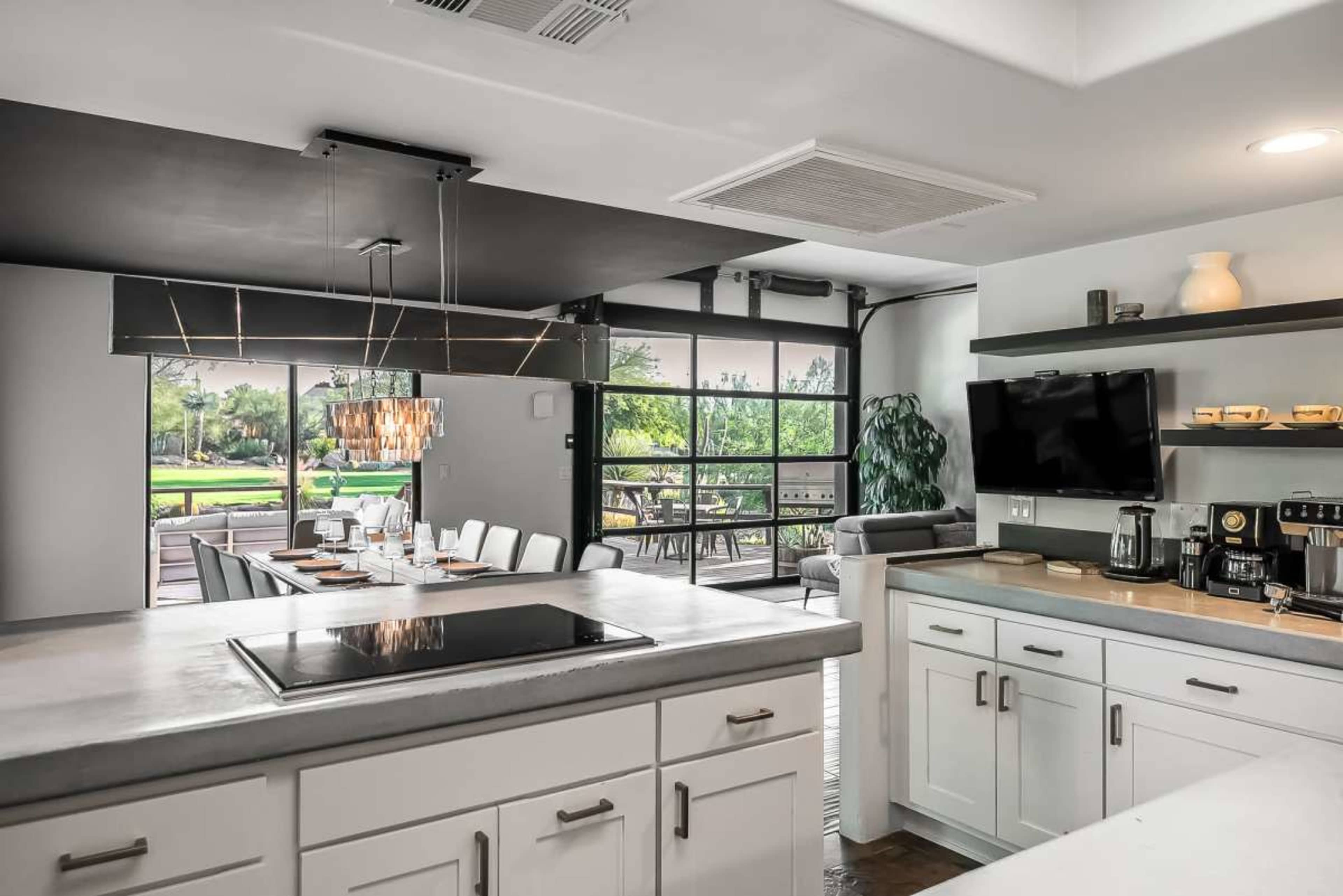 The image shows a modern kitchen with a concrete countertop, featuring an open layout that connects to a dining area and a view of an outdoor space through large windows.